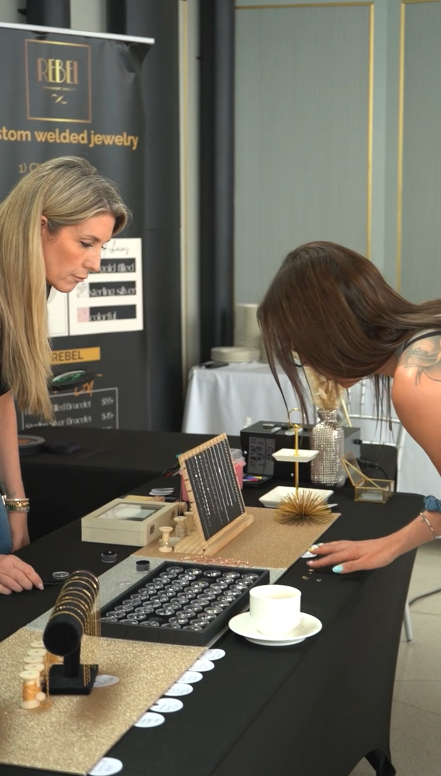 Two people browsing jewelry on a black table at a booth with displays and bracelets
