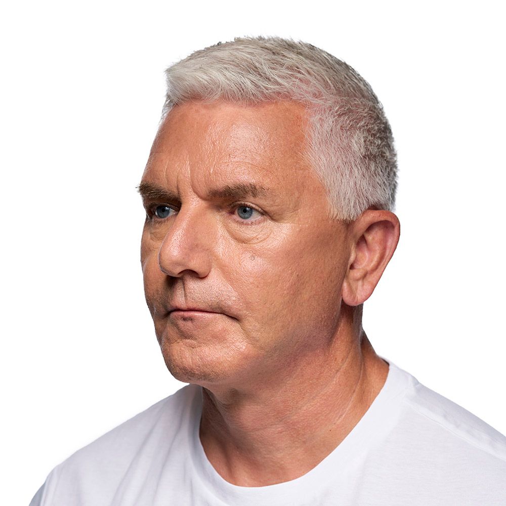 Man with short gray hair, wearing a white t-shirt, looking to the left with a serious expression.