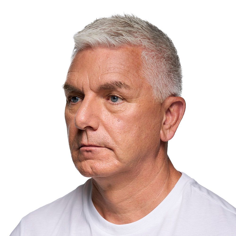 Man with gray hair, wearing a white shirt, looking to the left with a neutral expression.