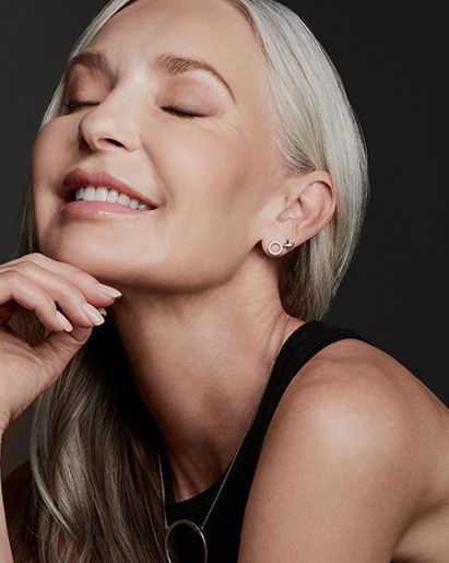 Woman with closed eyes and a smile, chin resting on hand, silver hair, black top.