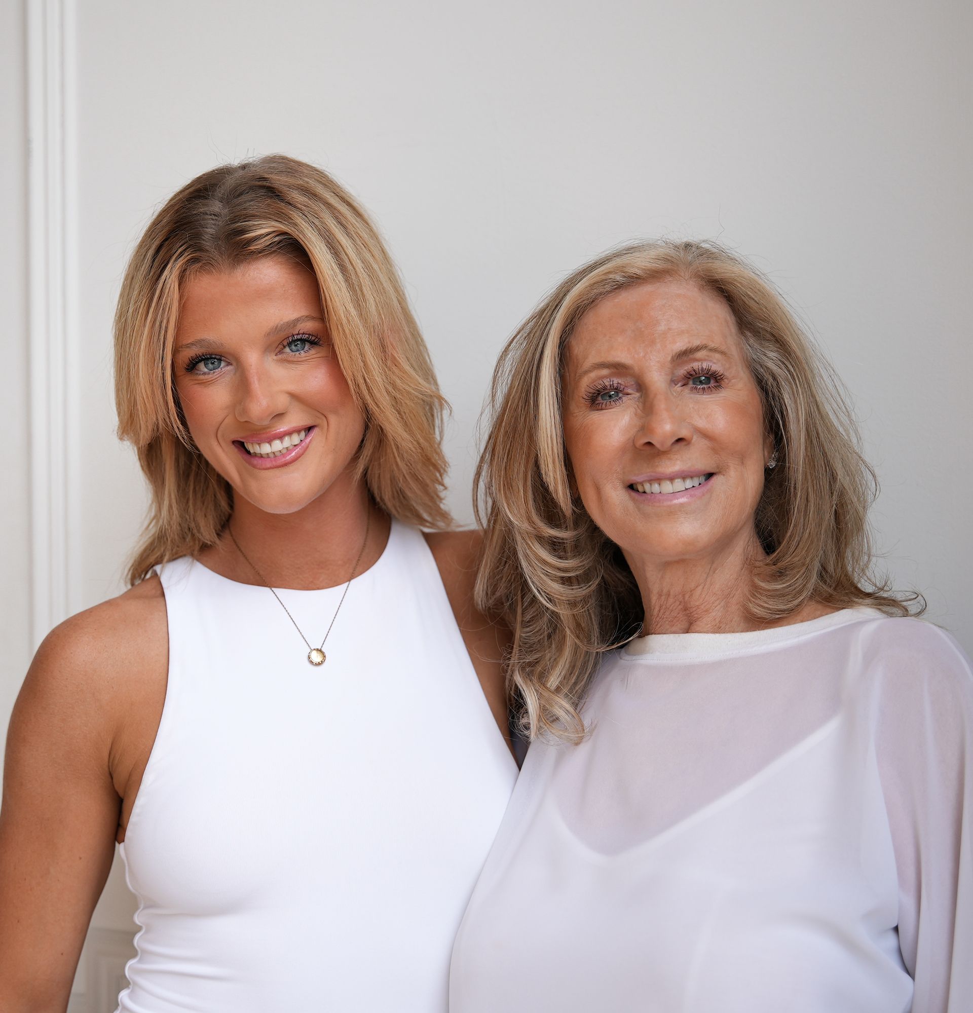 Young woman and older woman, both with blonde hair, smiling and posing indoors. They wear white tops.