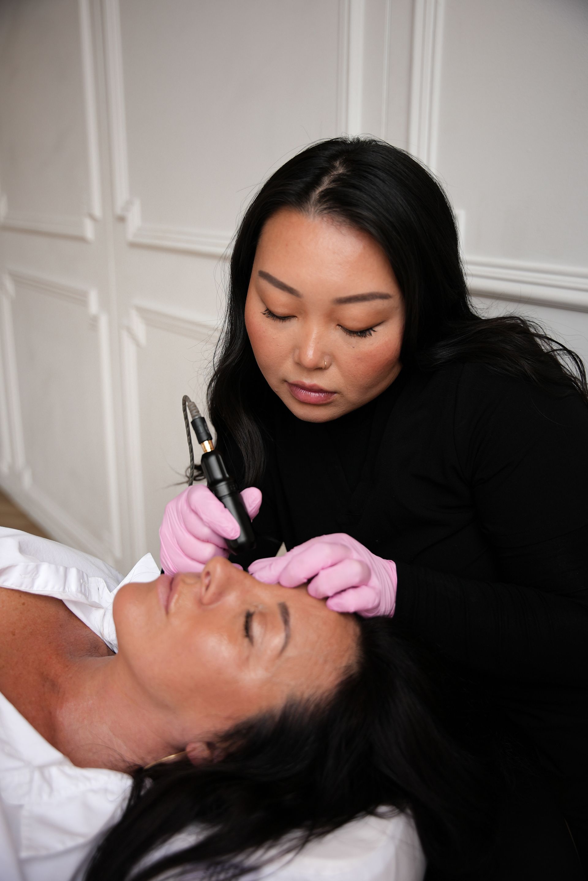 Woman receiving eyebrow microblading; technician in black, wearing pink gloves; white room.