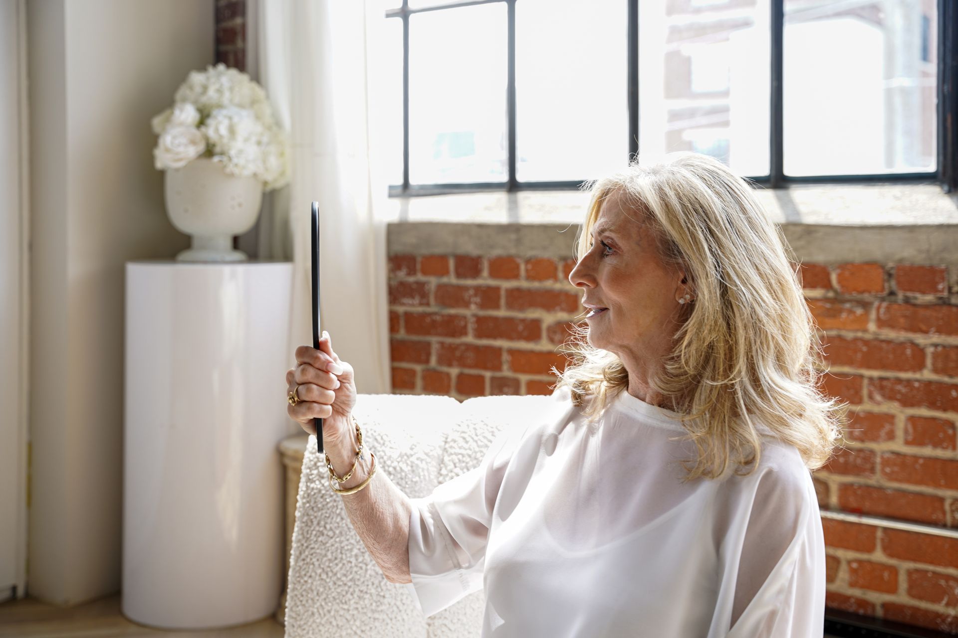 Blonde woman smiles while looking into a handheld mirror, near a window and red brick wall.