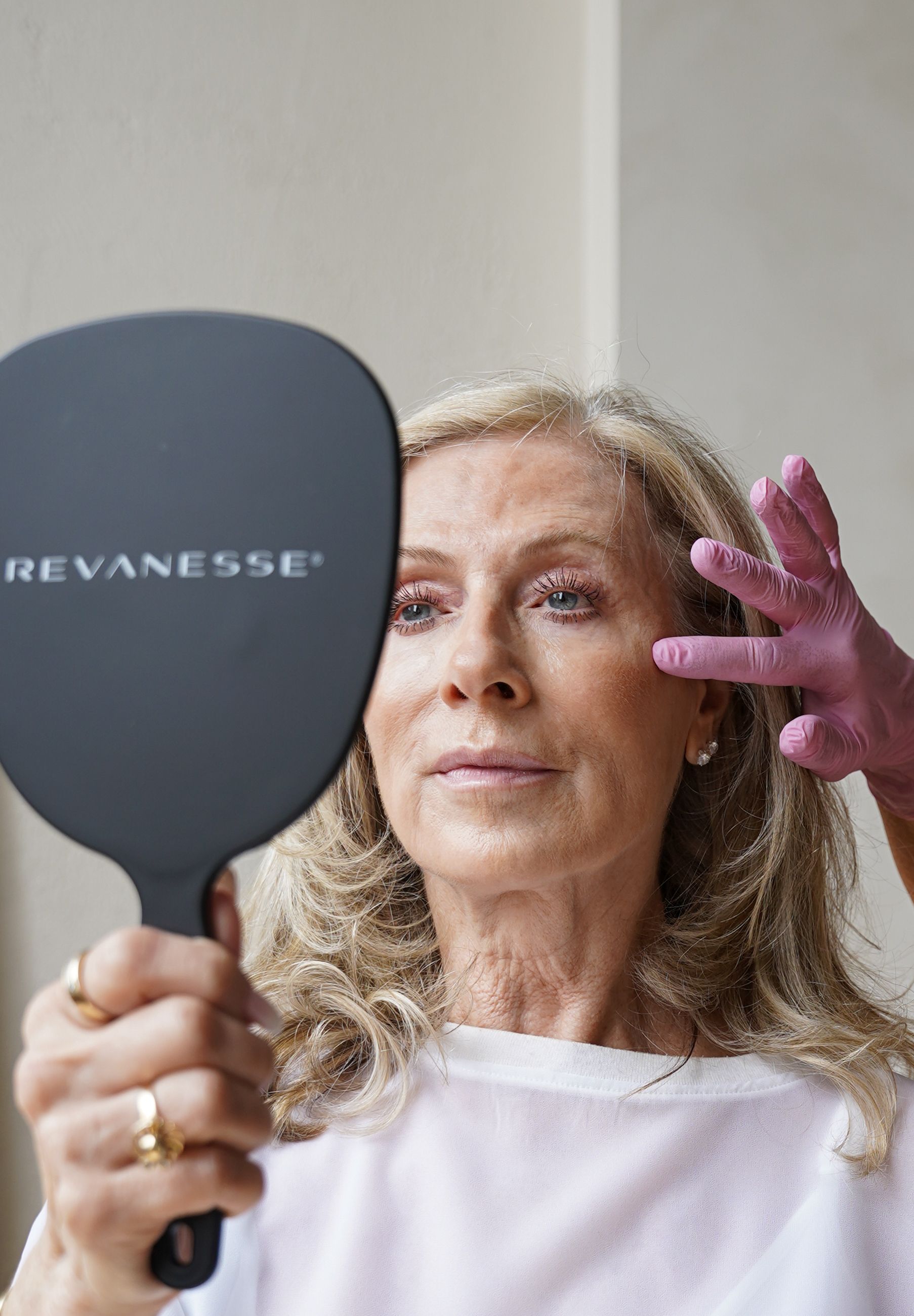 Woman applies pink makeup while holding a black REVANESSE mirror in front of a light wall.