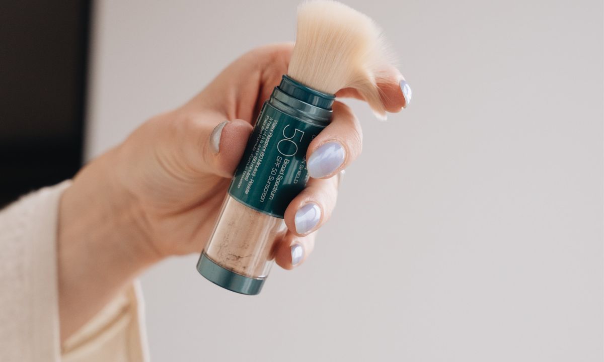 Hand holding a teal cosmetic stick with beige cream at the top against a blurred background