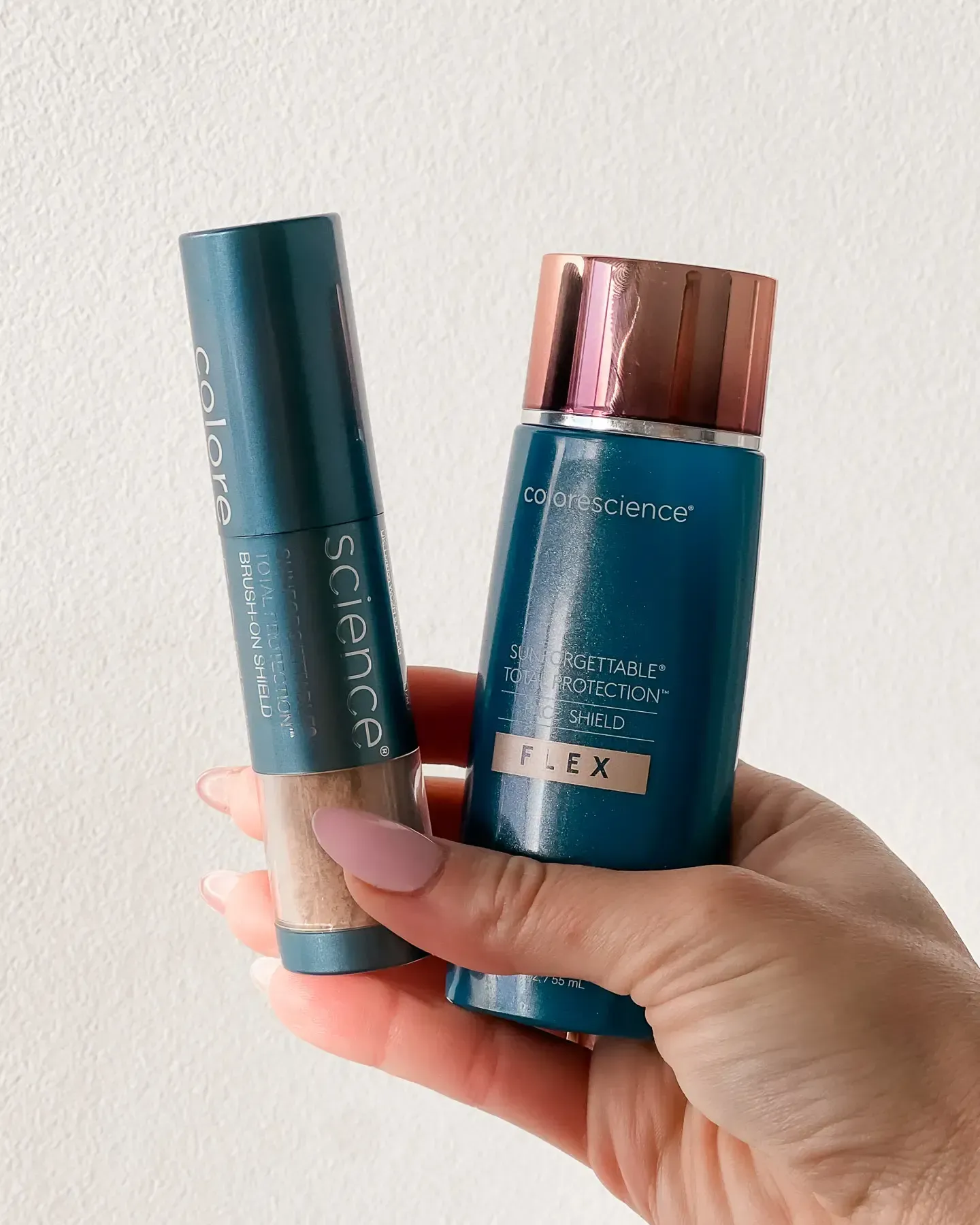 Hand holding two teal beauty bottles, one with a copper cap, against a white wall