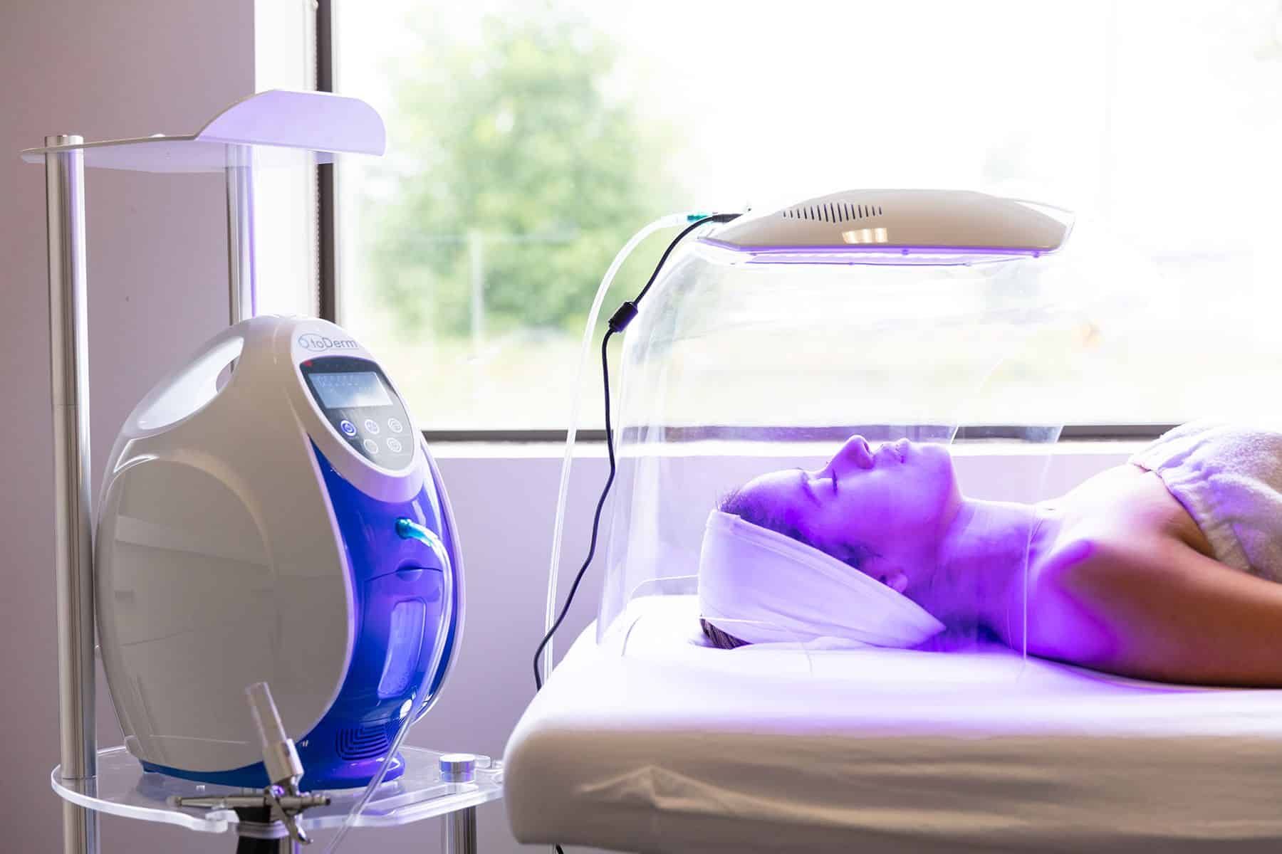 Woman receiving light therapy treatment, lying on a bed under a dome, illuminated by purple light.
