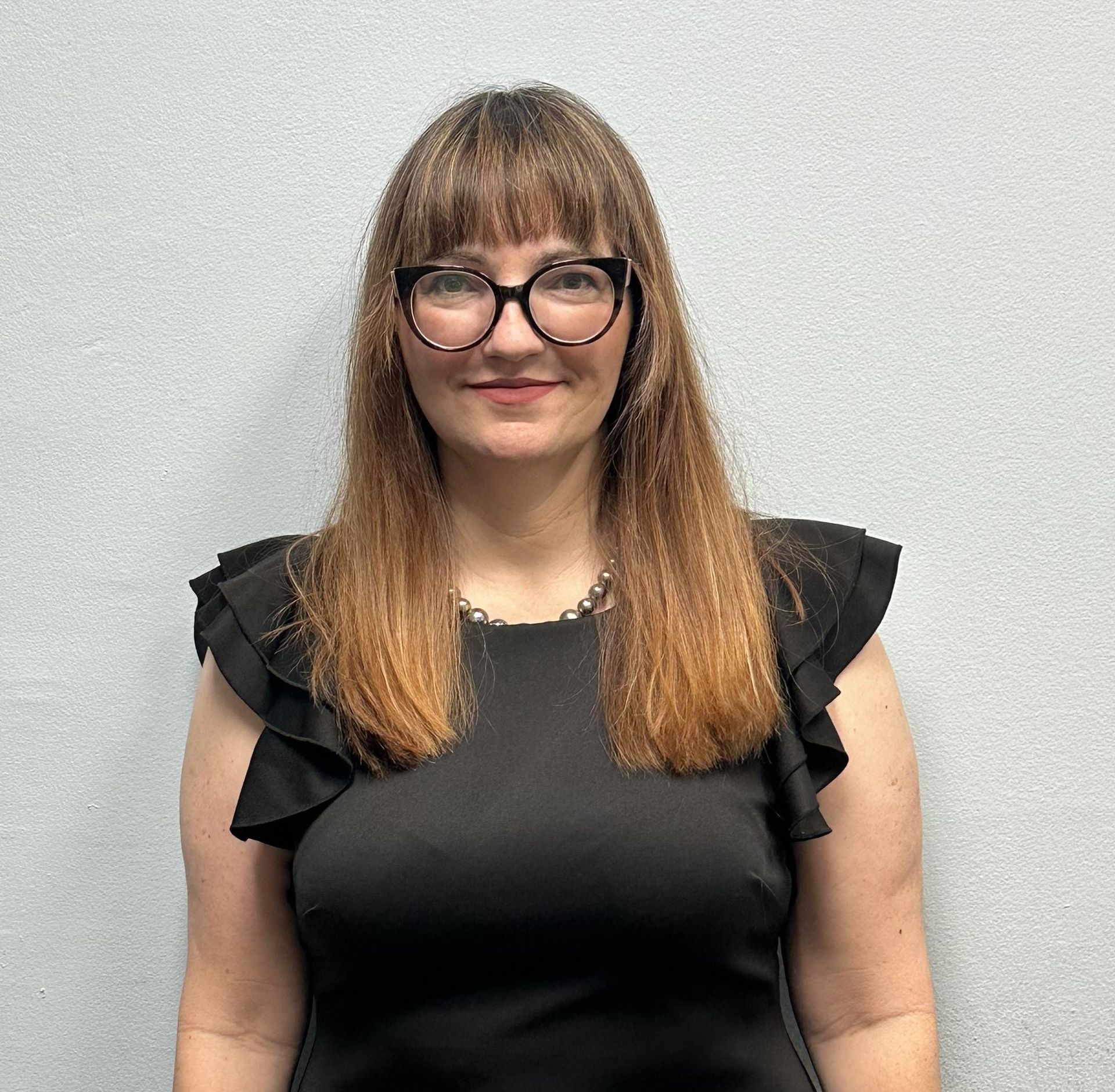 A woman wearing glasses and a black top is standing in front of a wall.