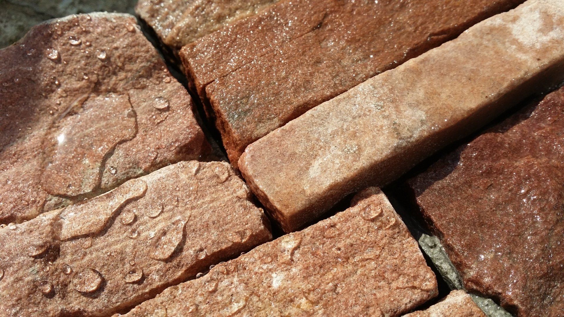 Zbliżenie na mokre, czerwonobrązowe, prostokątne cegły o różnej wielkości i fakturze.