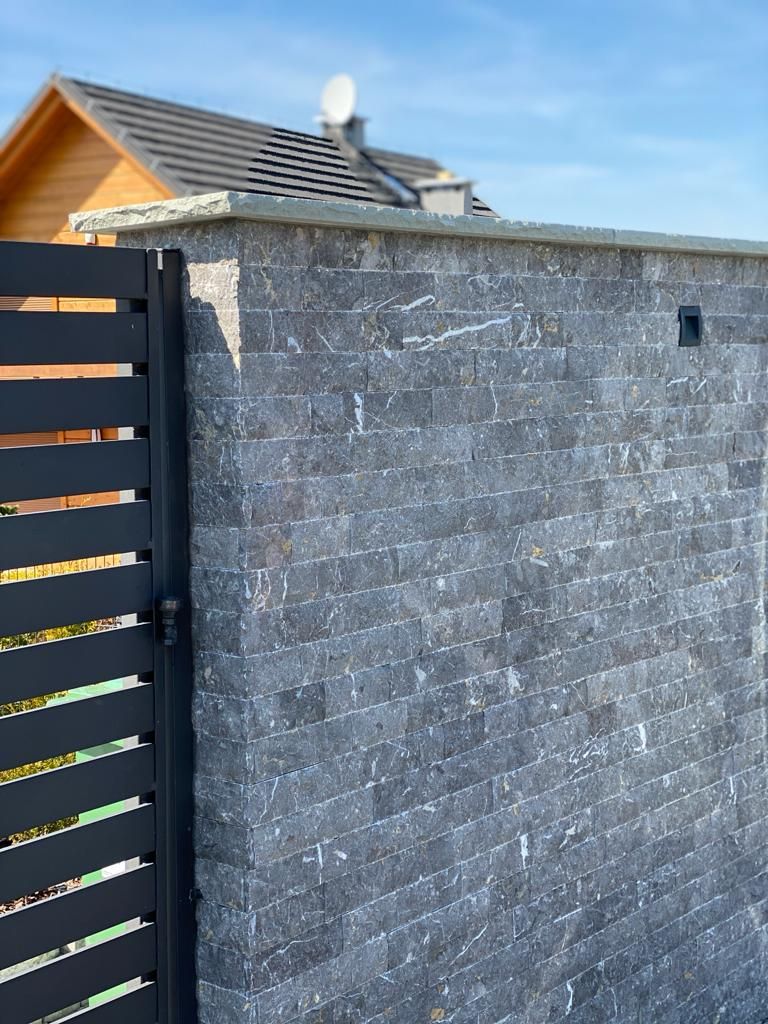 Kamienny mur z czarną metalową bramą; w tle widoczny jest drewniany dom z ciemnym dachem.