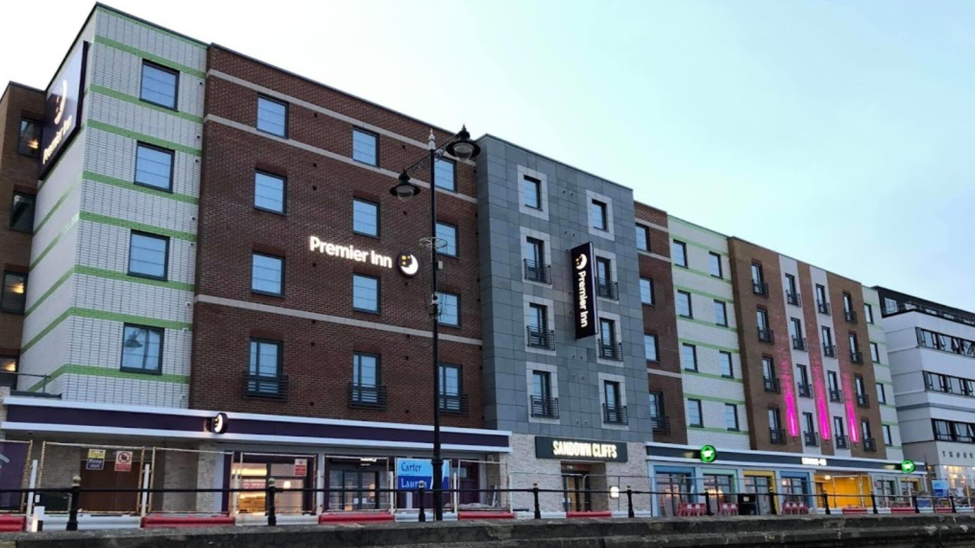 Zewnętrzna część hotelu Premier Inn z różnokolorowymi fragmentami cegły, oknami i witrynami sklepowymi poniżej.