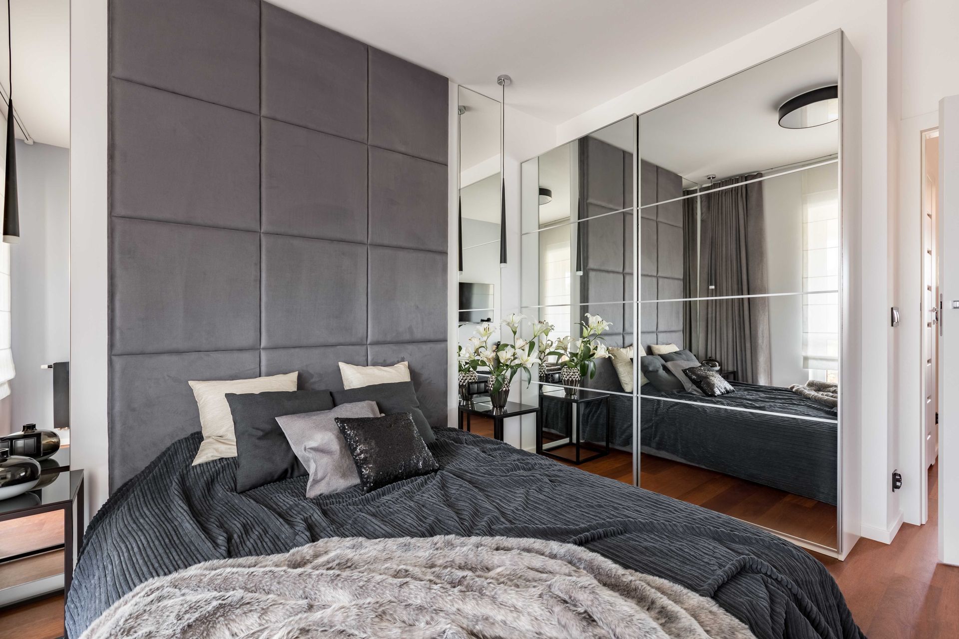 Chambre moderne avec literie gris foncé, portes de placard à miroir et grande tête de lit capitonnée grise.