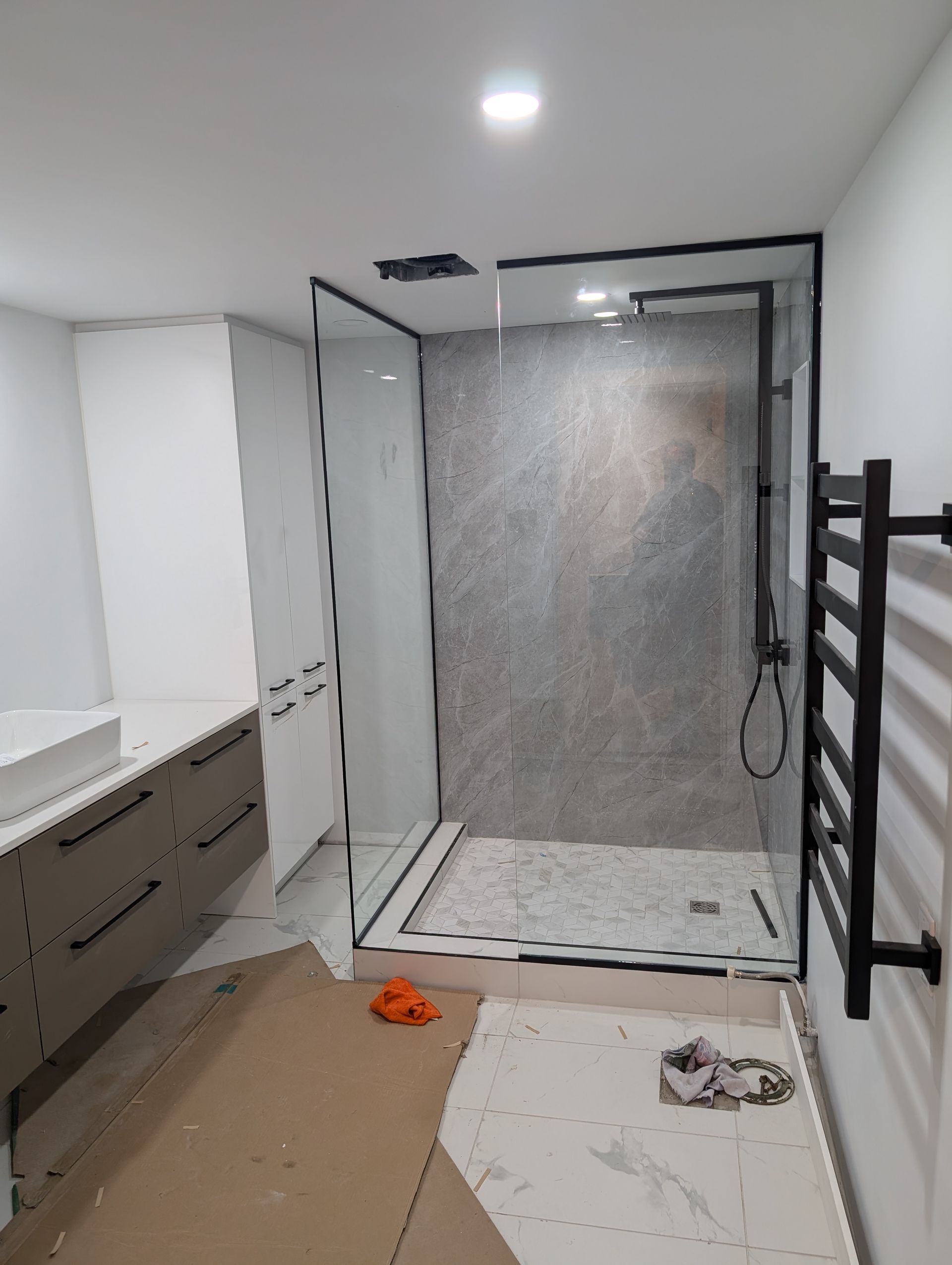 Salle de bains avec espace douche, meuble-lavabo blanc et porte-serviettes noir ; le sol semble inachevé et jonché d’objets.