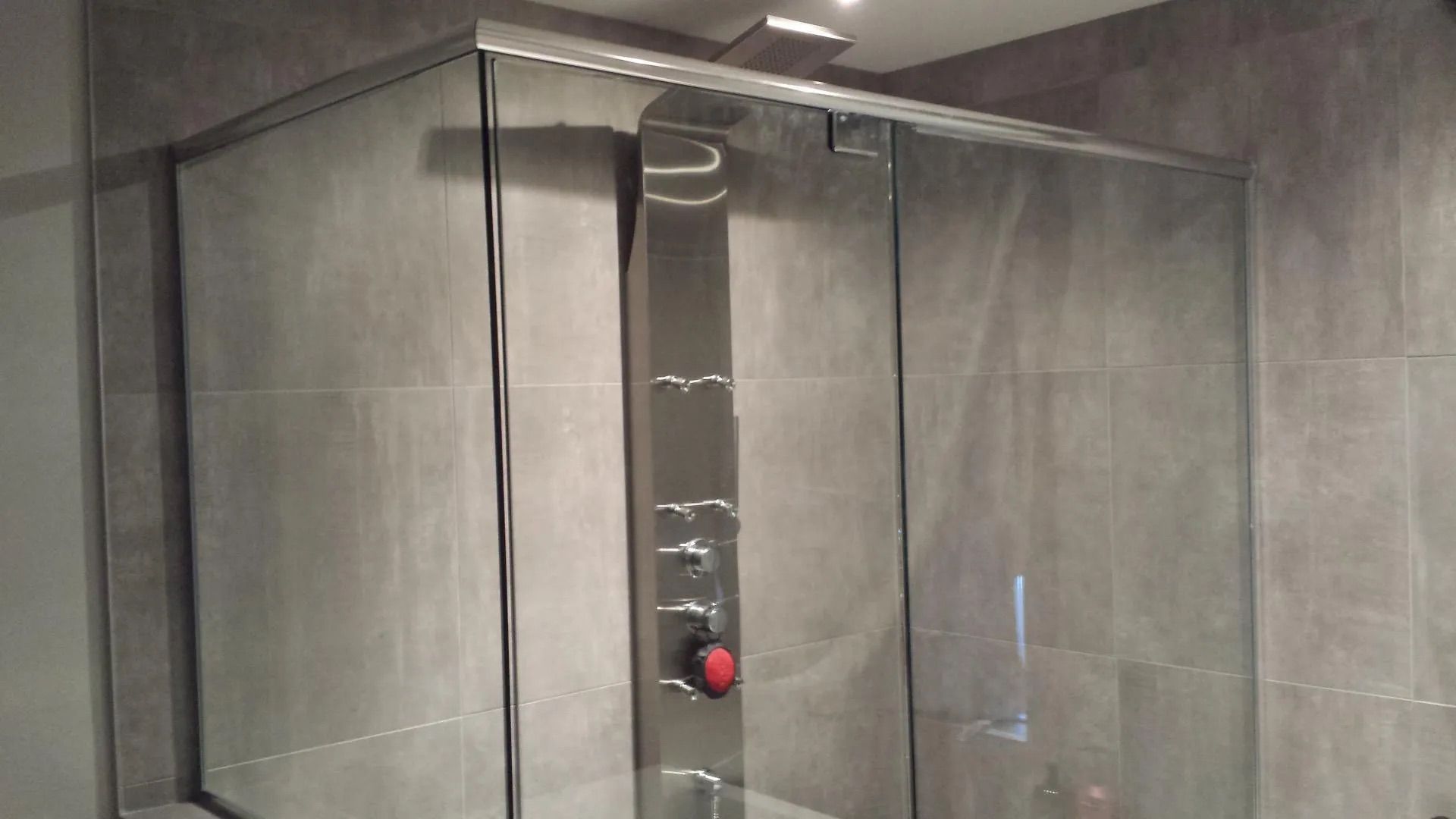 Cabine de douche en verre dans une salle de bains carrelée de gris avec une commande de douche rouge visible