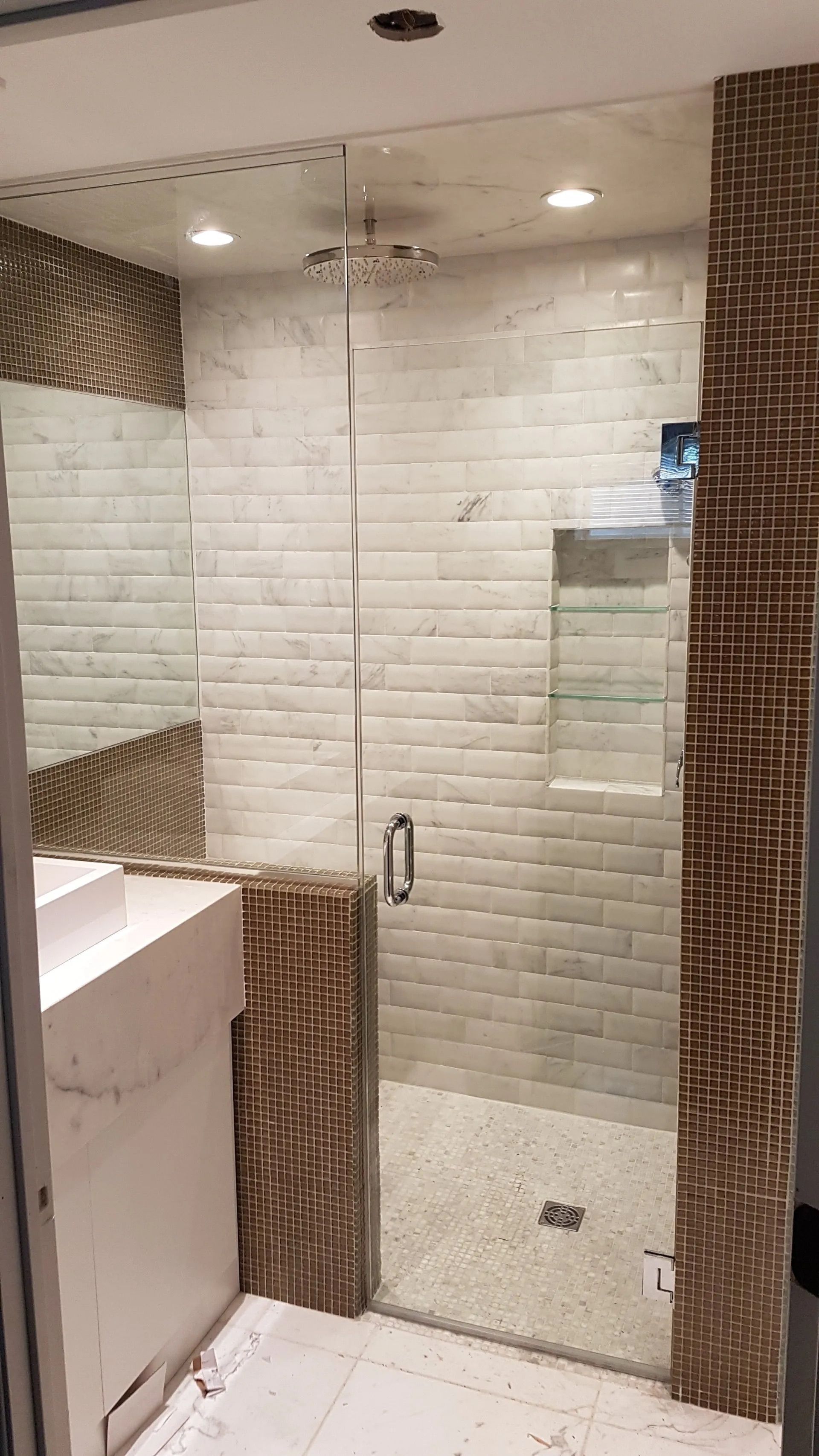 Douche moderne avec carrelage blanc, porte en verre, carreaux décoratifs marron et lavabo sur le côté gauche.
