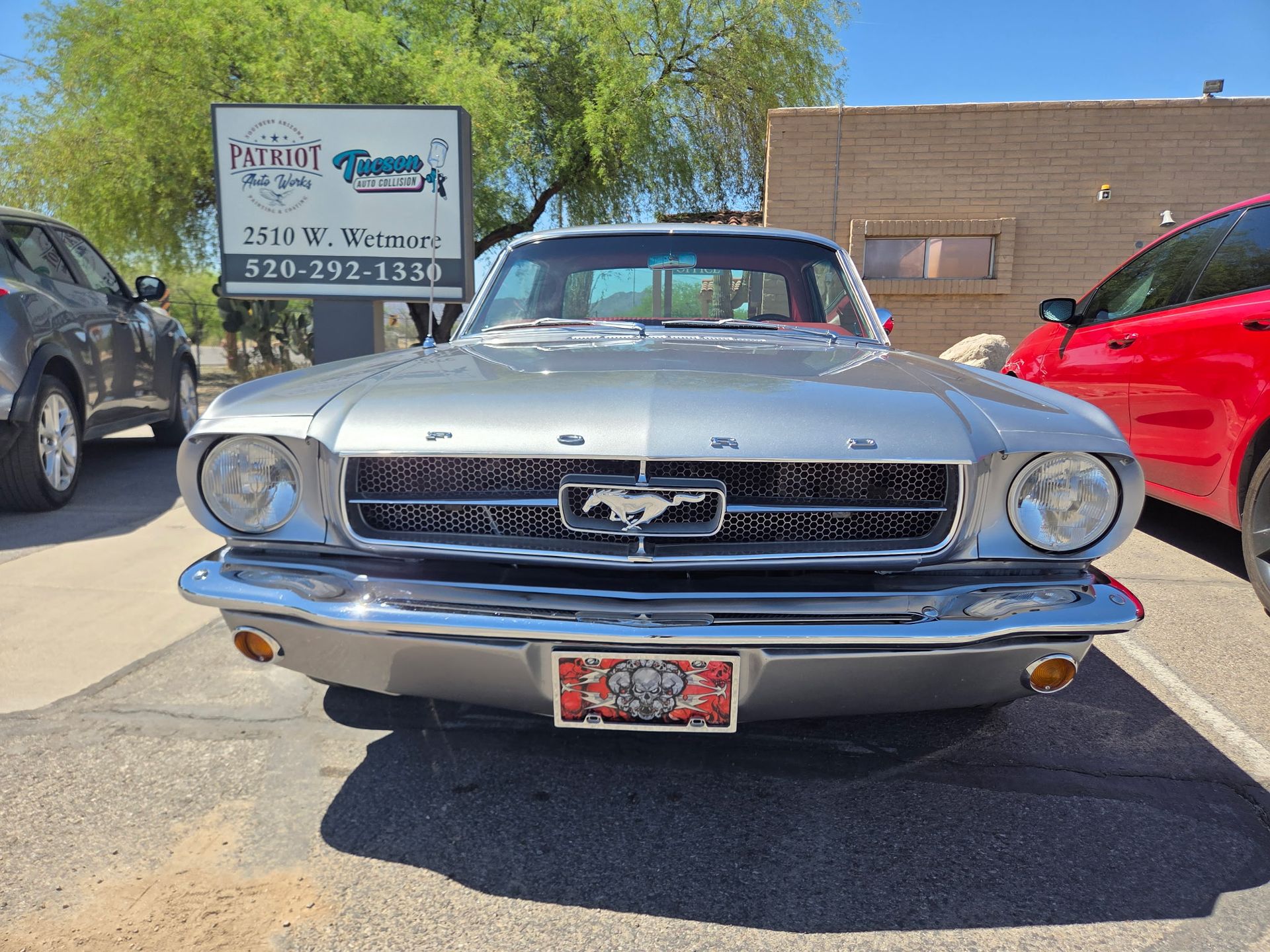 Front view of a silver classic Ford Mustang parked outdoors beside other cars and a sign.