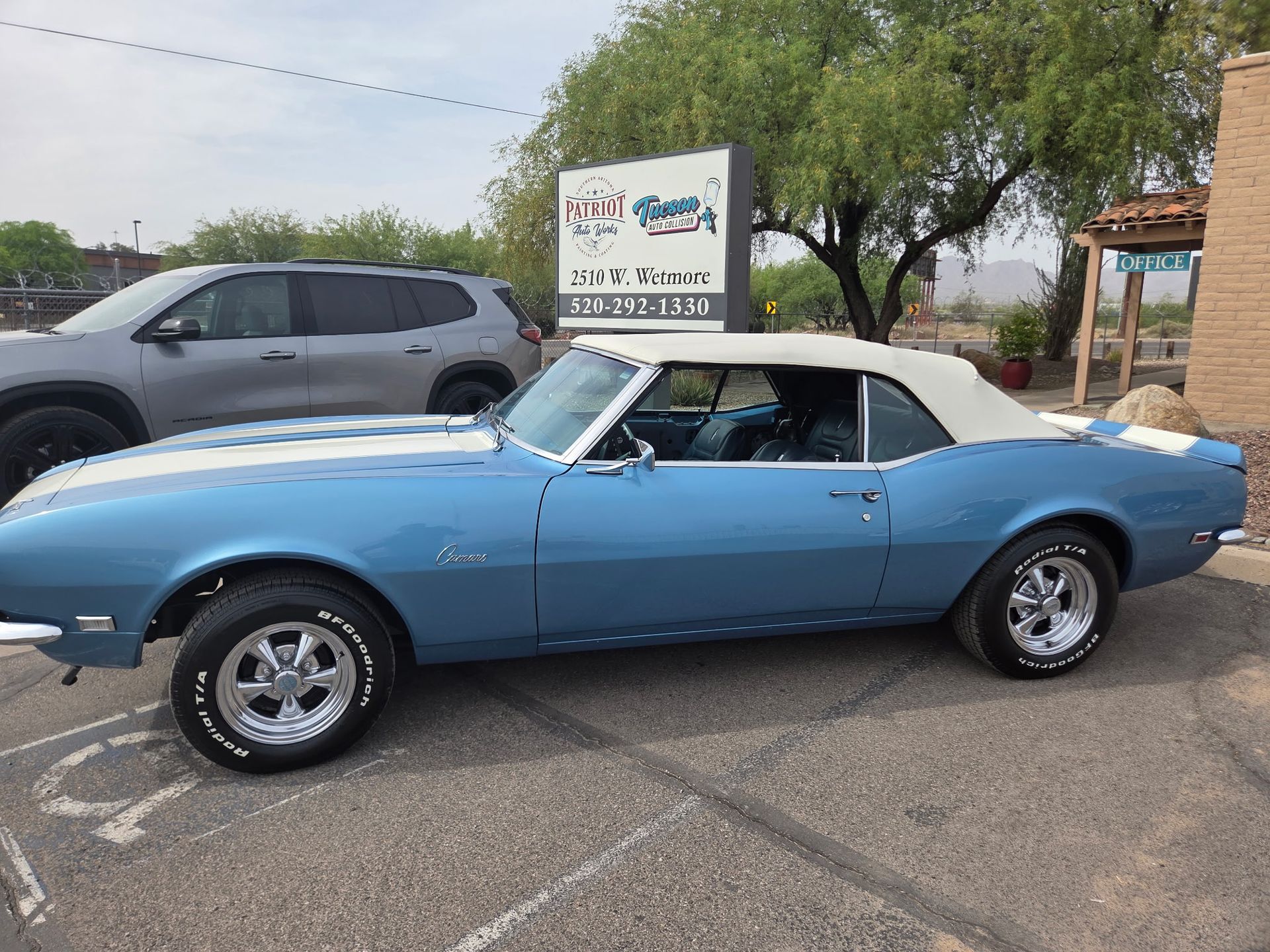 Blue classic convertible parked in a lot beside a gray SUV and a roadside sign.