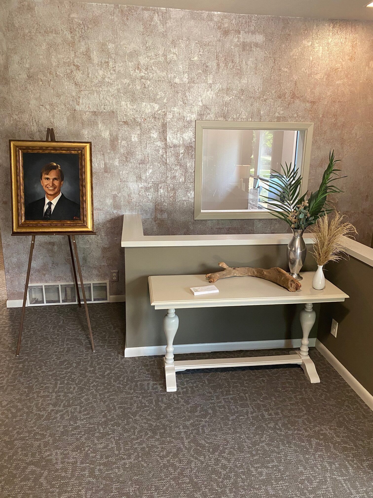 Framed portrait on easel beside a white table with flowers and mirror against a textured wall.