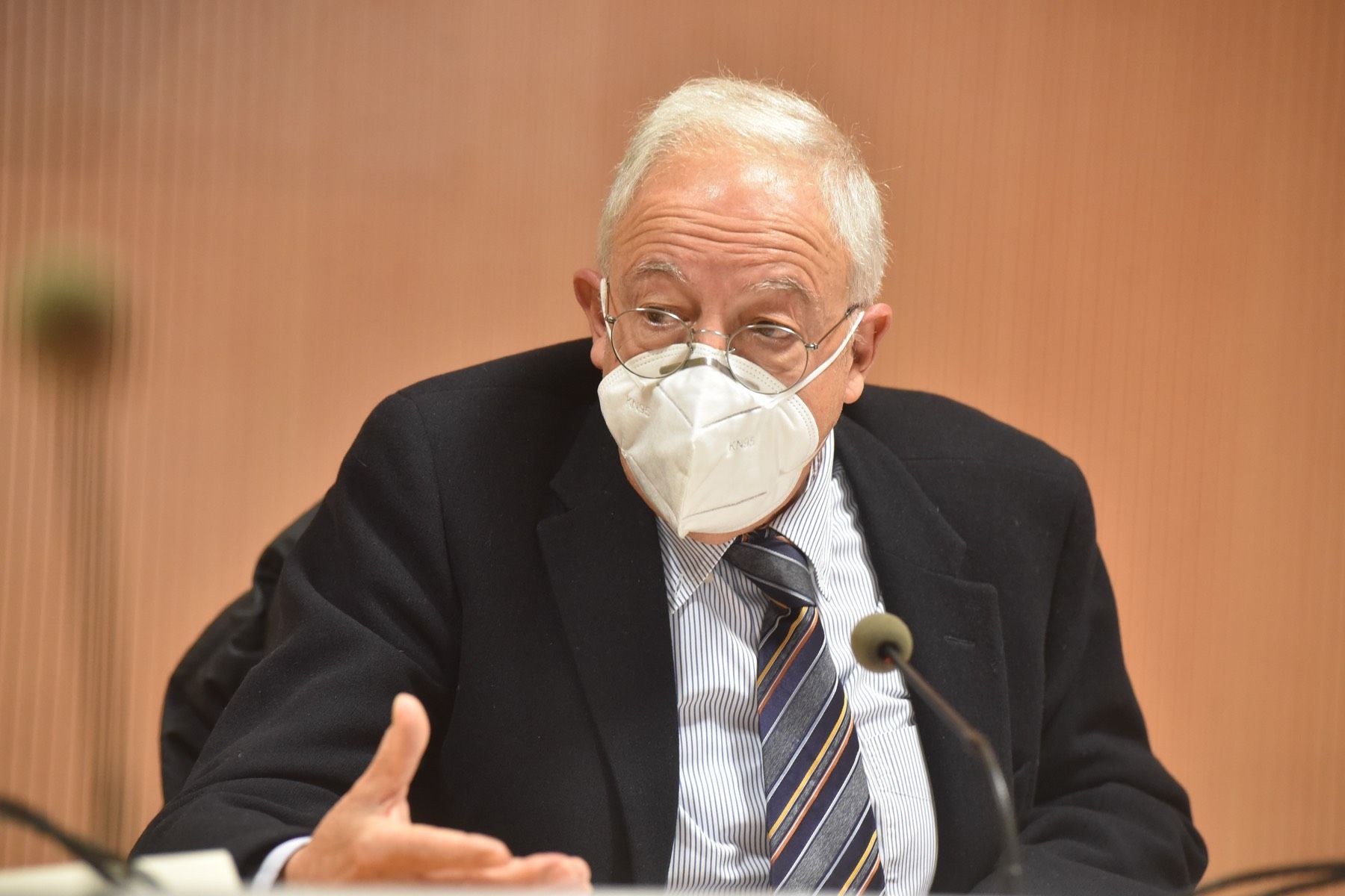 Un uomo in giacca e cravatta indossa una maschera e parla al microfono.