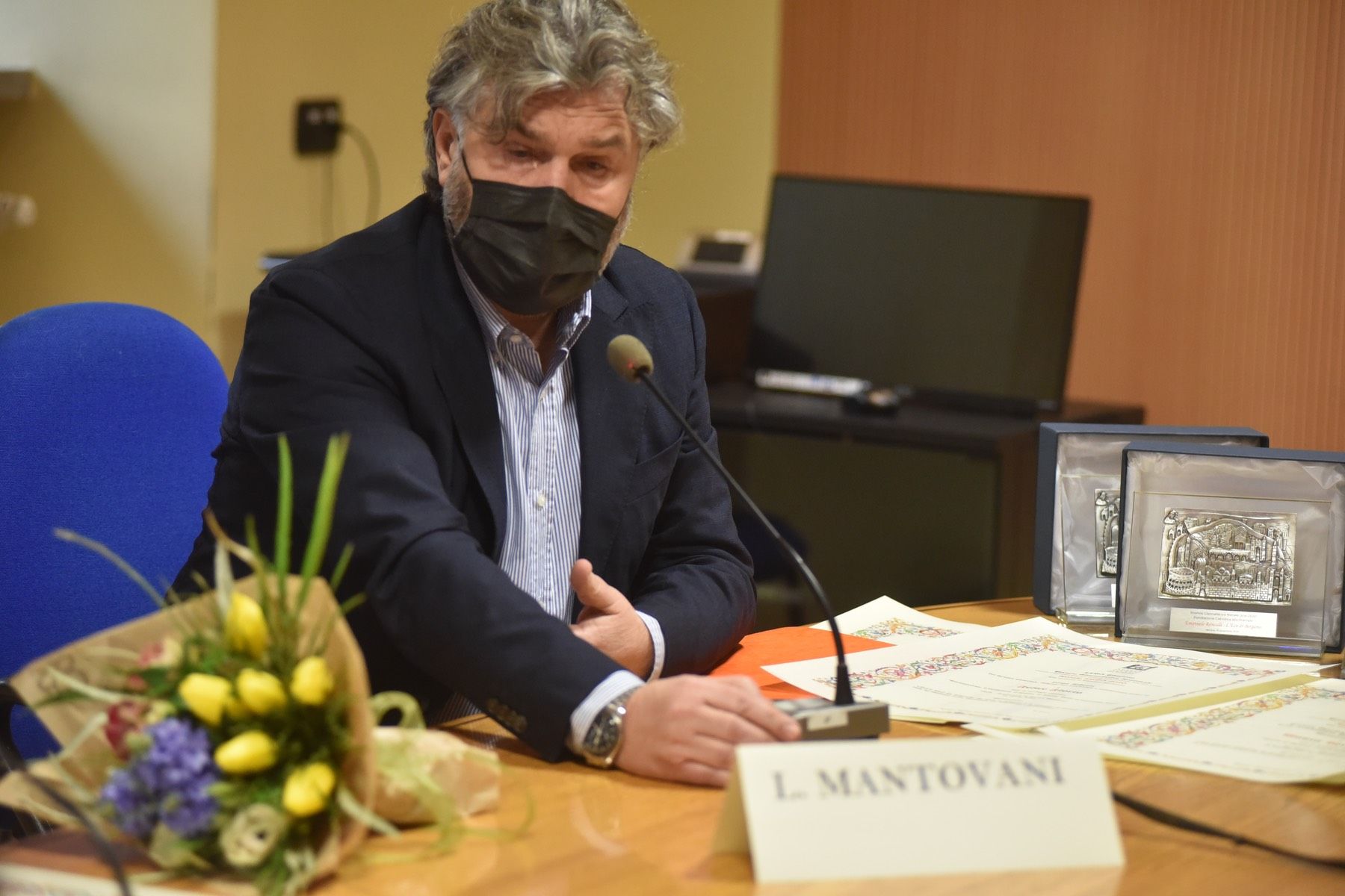 Un uomo che indossa una maschera è seduto a un tavolo con fiori e un microfono.