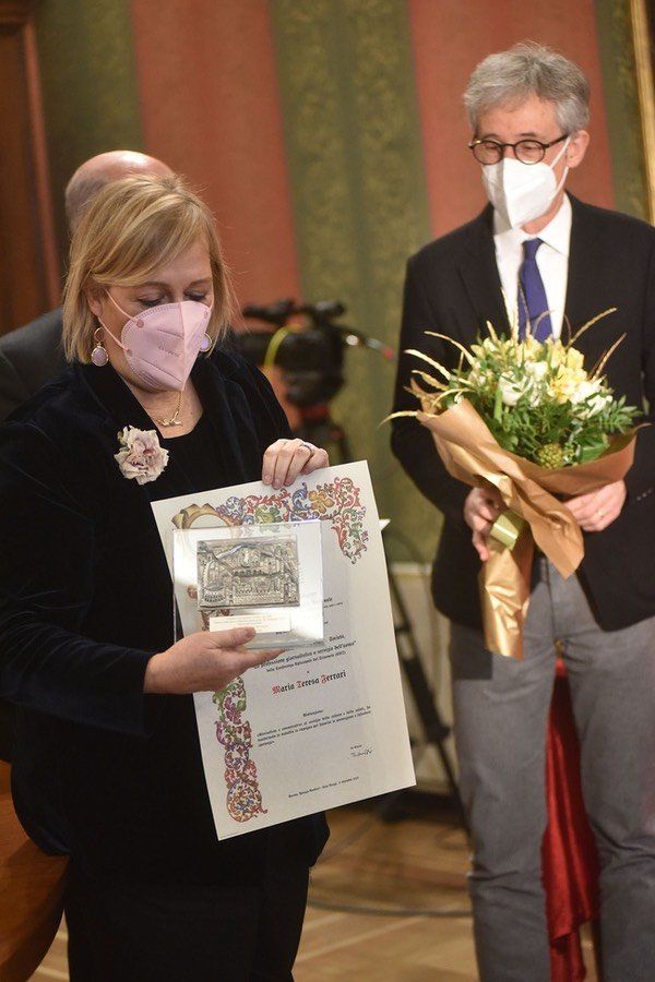 Una donna che indossa una maschera tiene in mano un certificato e un mazzo di fiori.