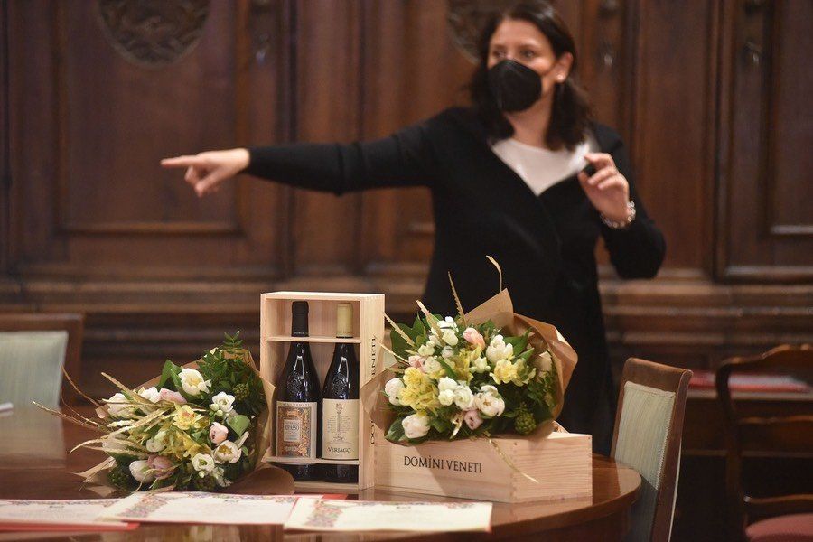 Una donna che indossa una maschera è in piedi davanti a un tavolo con fiori e bottiglie di vino.