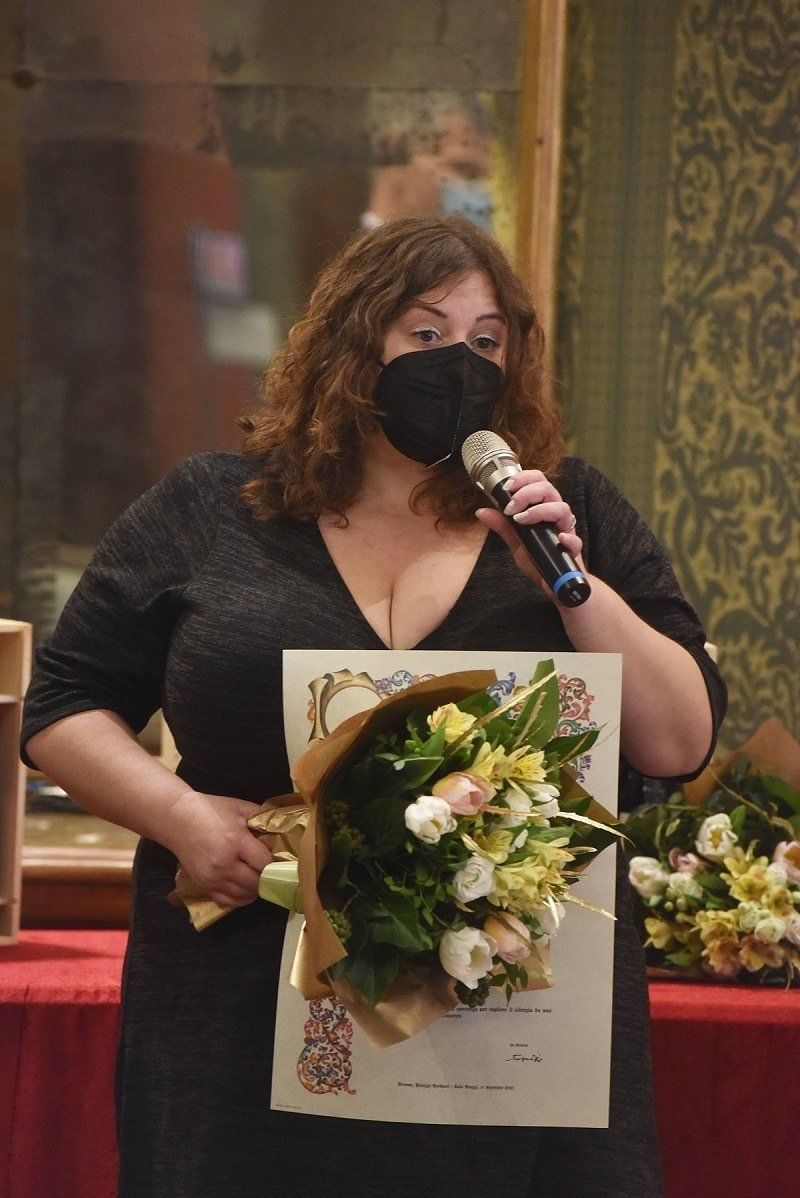 Una donna che indossa una maschera tiene in mano un mazzo di fiori e un certificato.