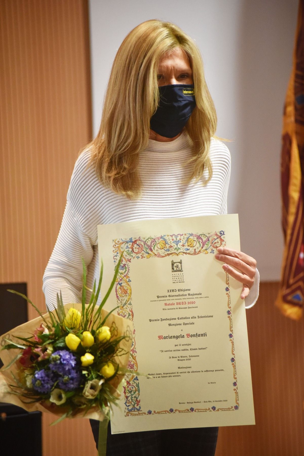 Una donna che indossa una maschera tiene in mano un certificato e un mazzo di fiori.