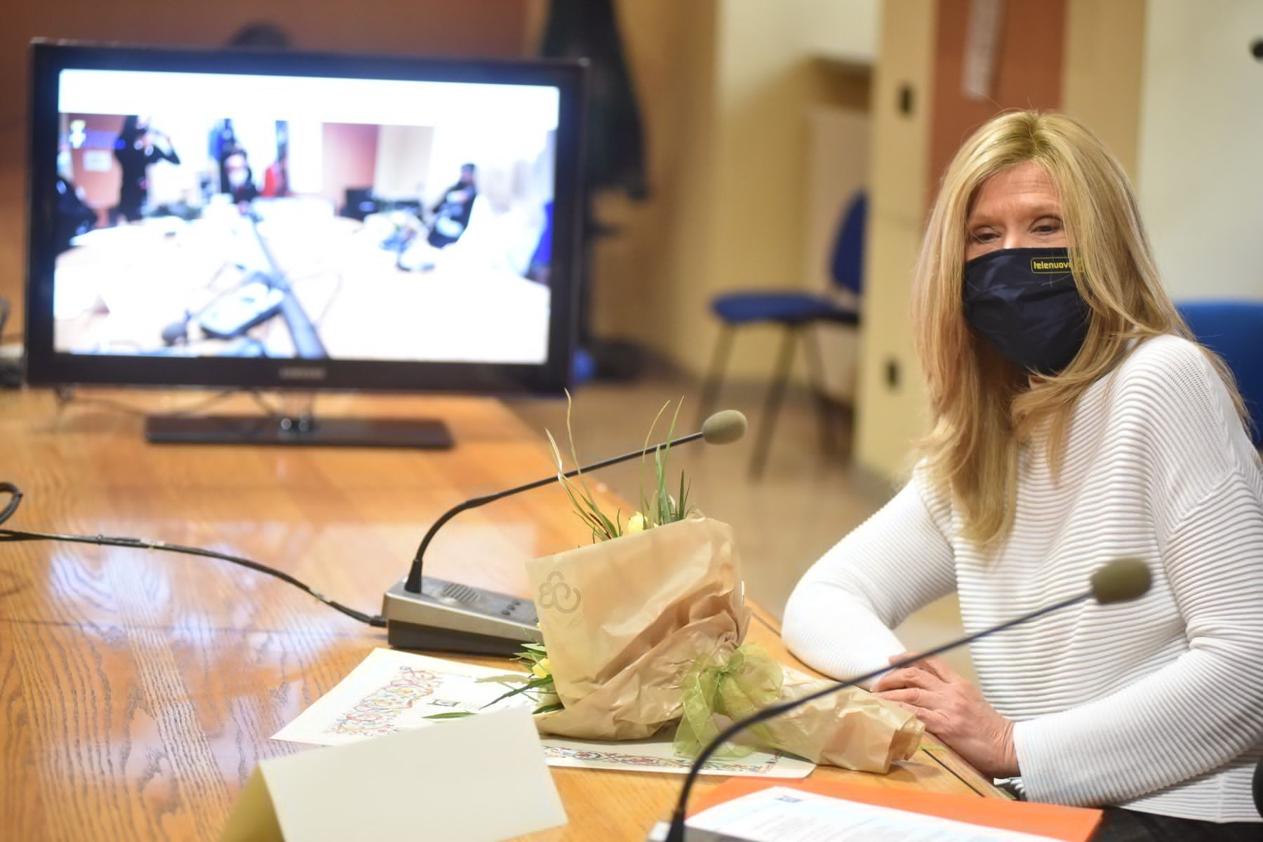 Una donna che indossa una maschera è seduta a un tavolo con dei microfoni davanti a un televisore.