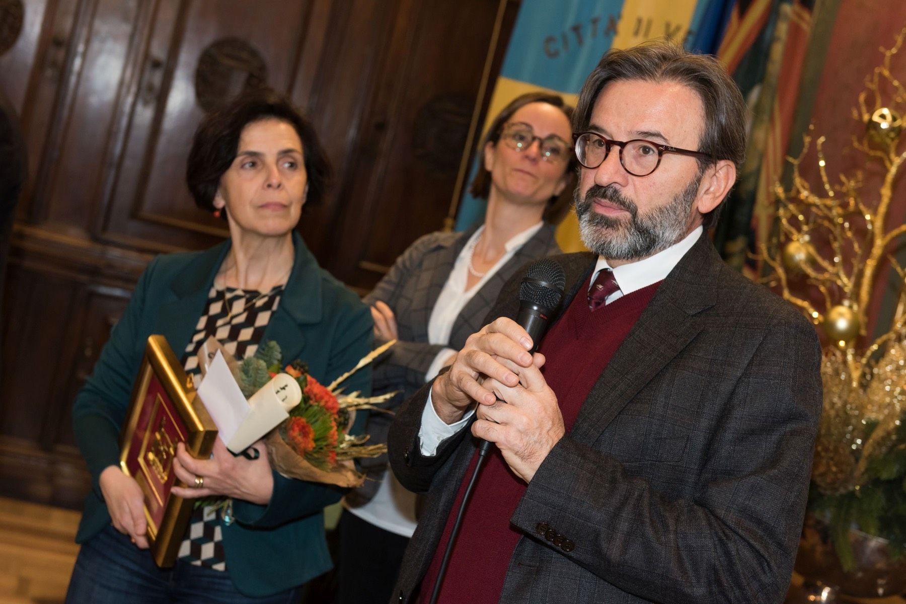 Un uomo con la barba parla al microfono mentre due donne stanno a guardare.