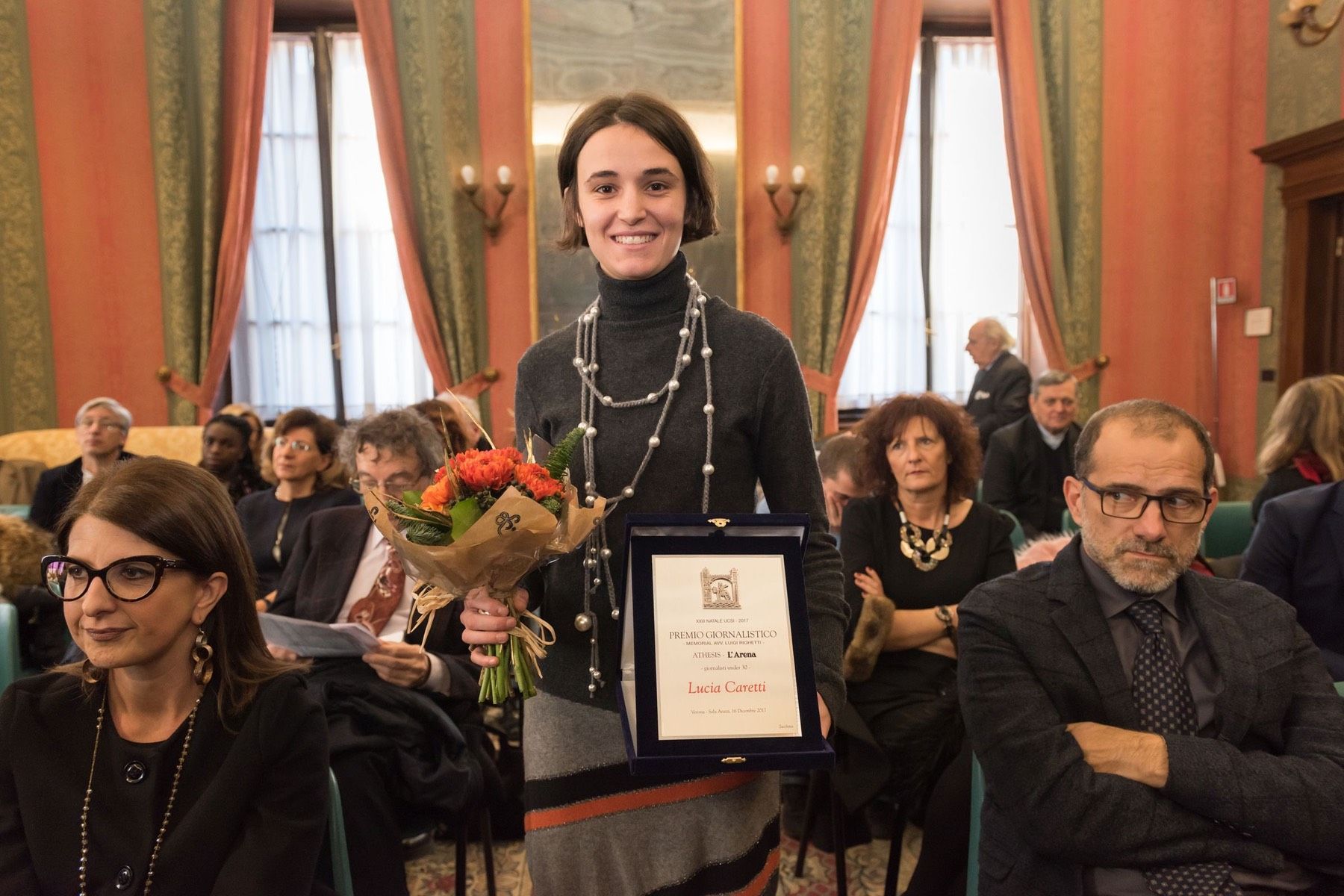 Una donna tiene in mano un mazzo di fiori e un certificato davanti a una folla di persone.