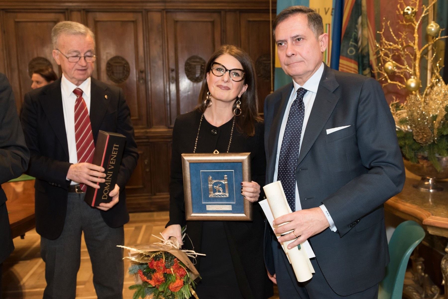 Una donna tiene in mano una foto incorniciata mentre sta in piedi accanto a due uomini in giacca e cravatta.