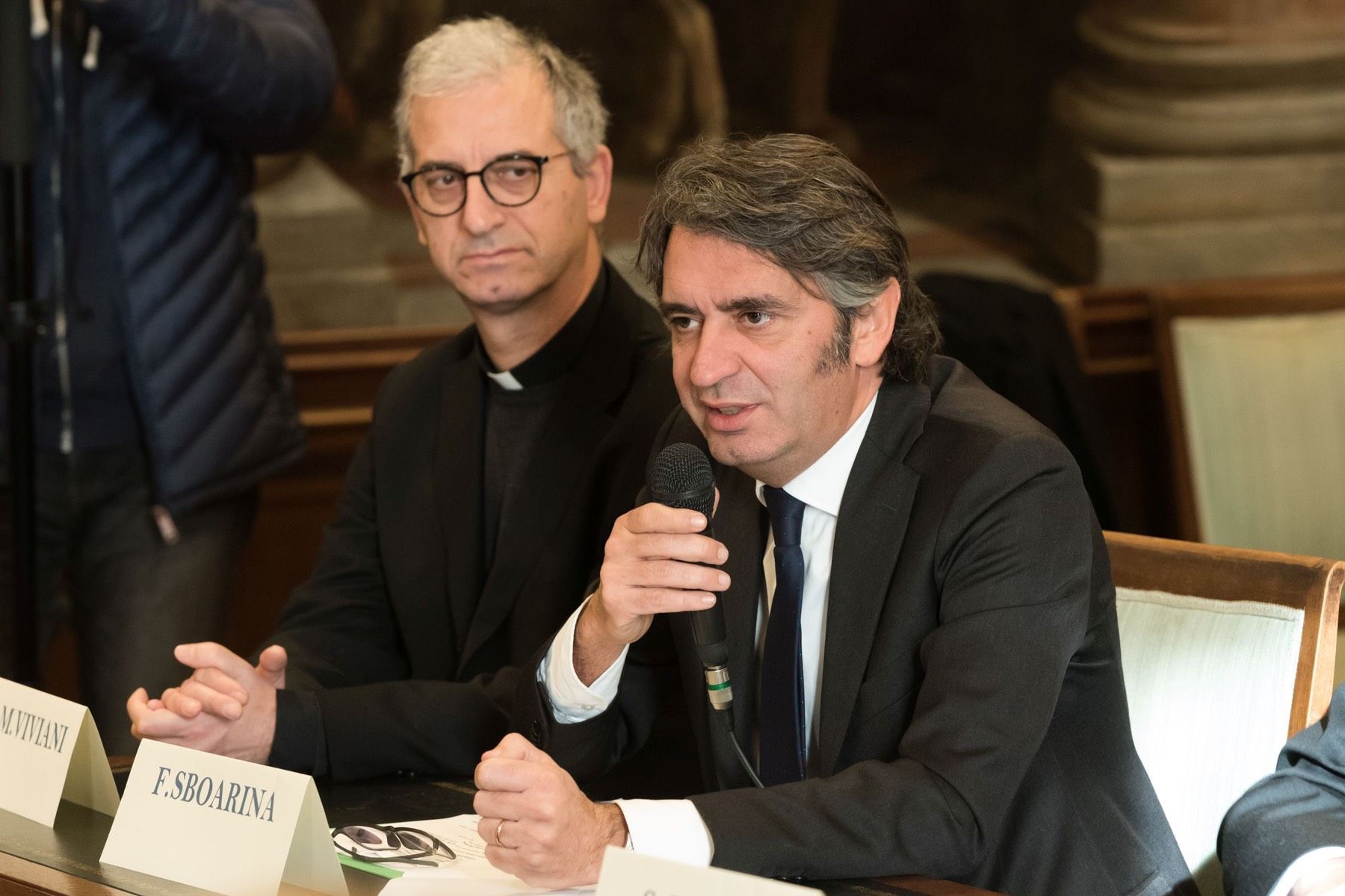 Due uomini sono seduti a un tavolo e parlano ai microfoni.