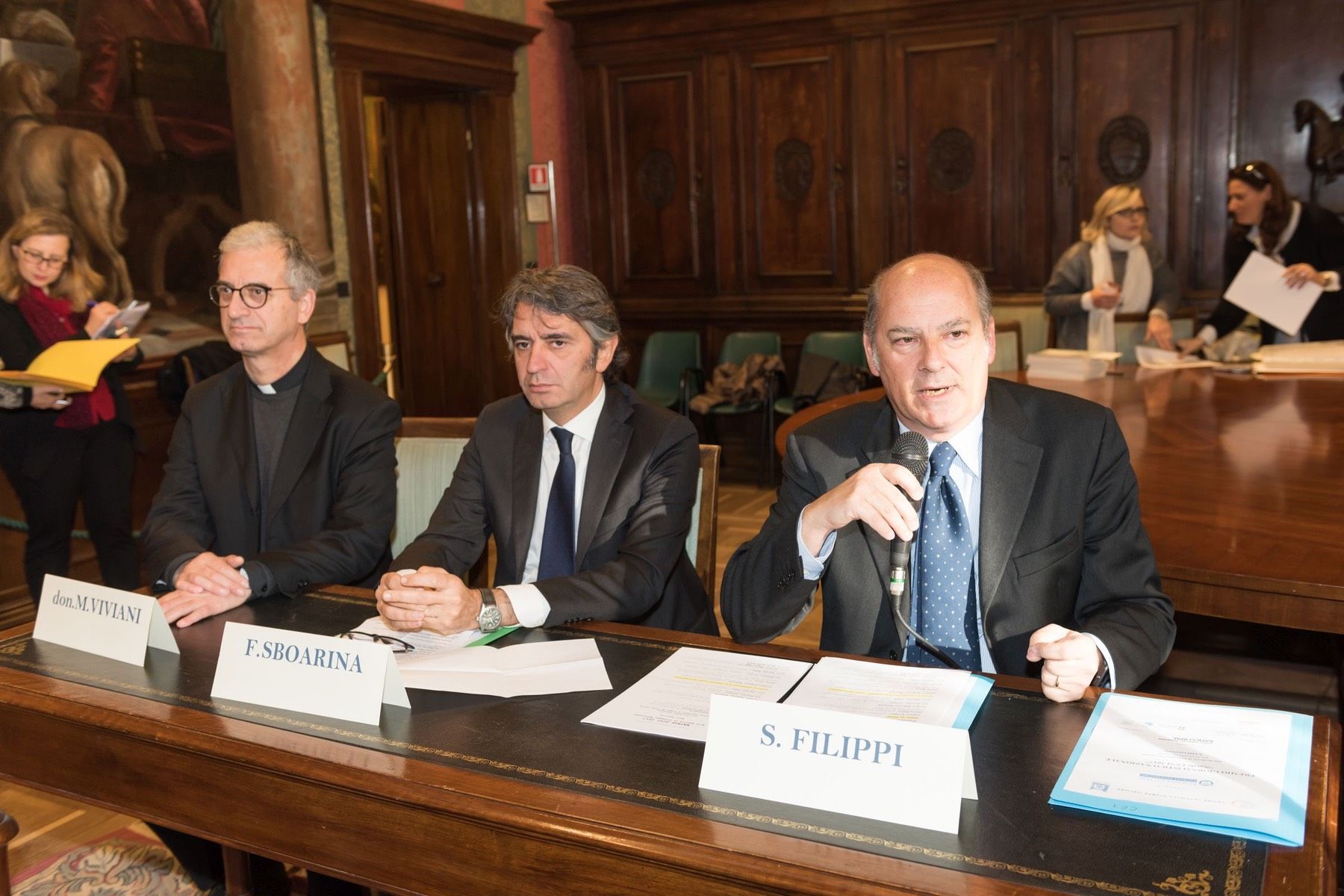 Un gruppo di uomini è seduto a un tavolo con dei documenti e un microfono.