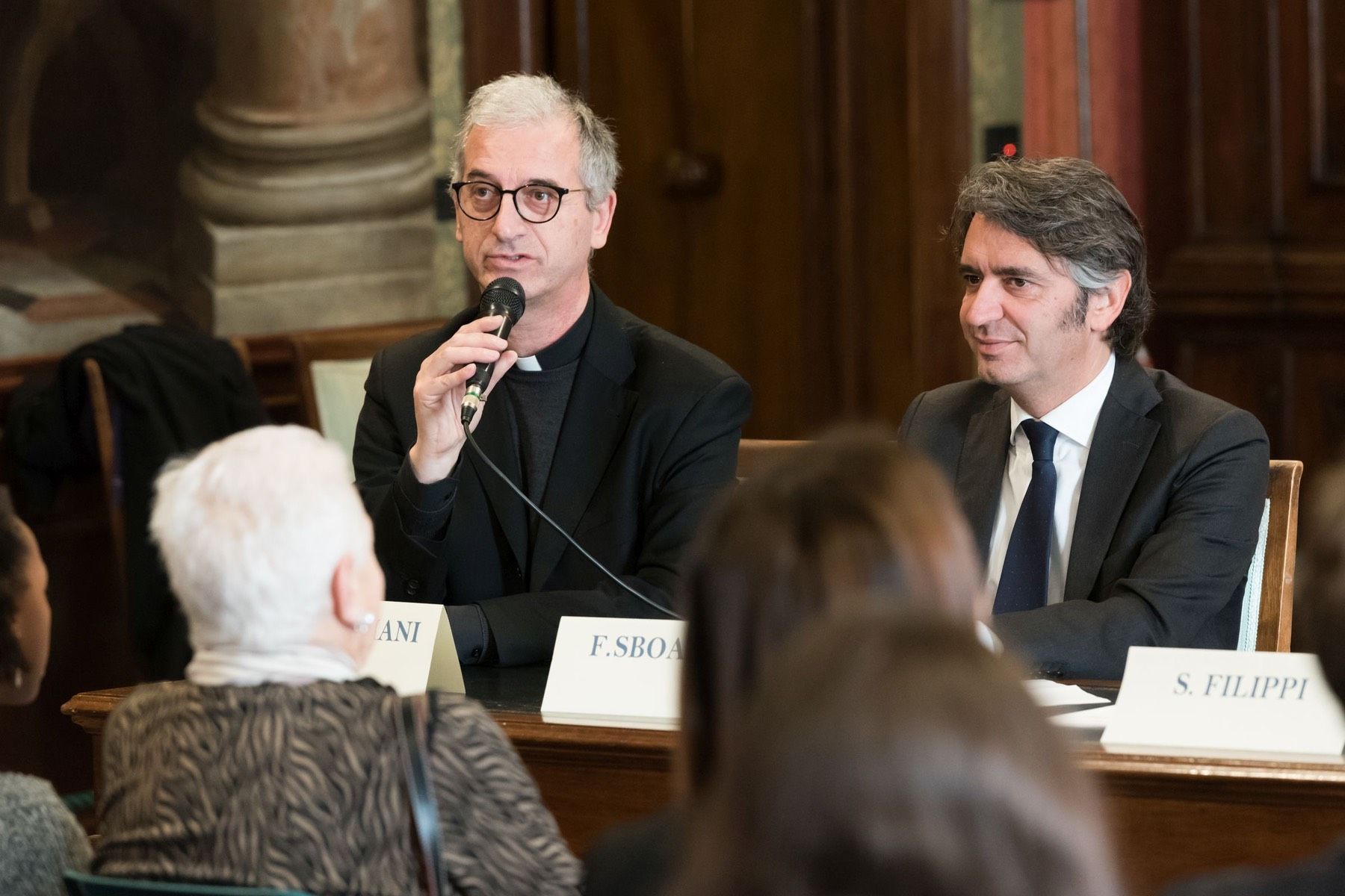 Due uomini sono seduti a un tavolo e parlano ai microfoni.
