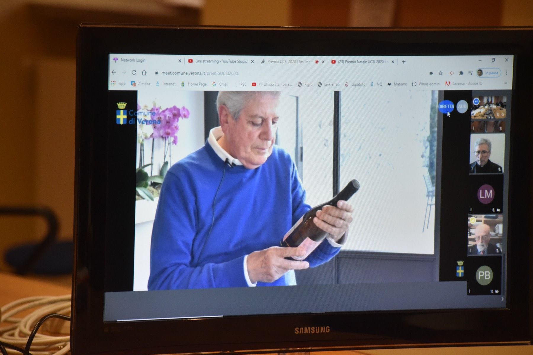 Un uomo con un maglione blu tiene in mano una bottiglia di vino sullo schermo di un computer.