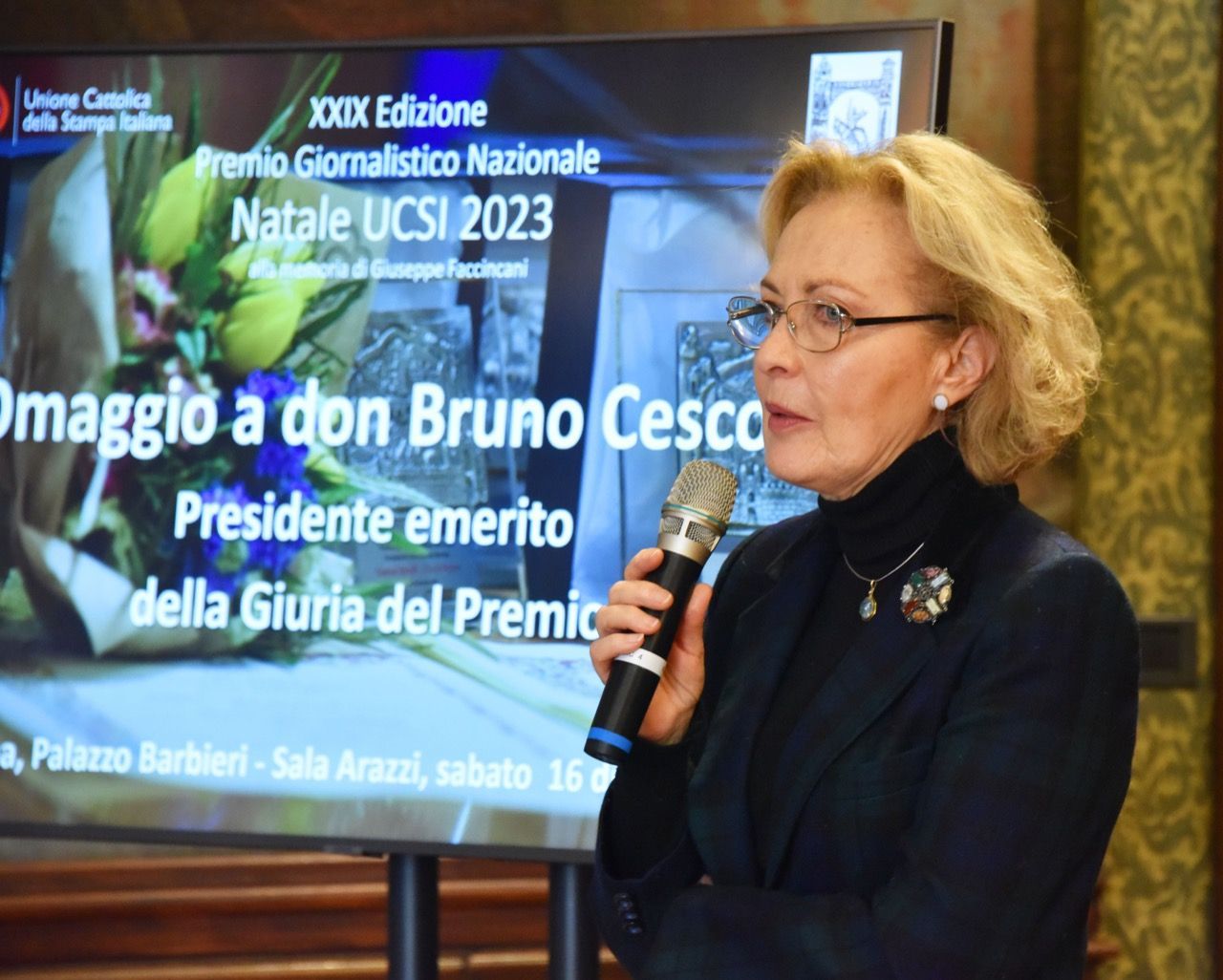 Una donna tiene in mano un microfono davanti a uno schermo su cui è scritto natale ucsi 2023