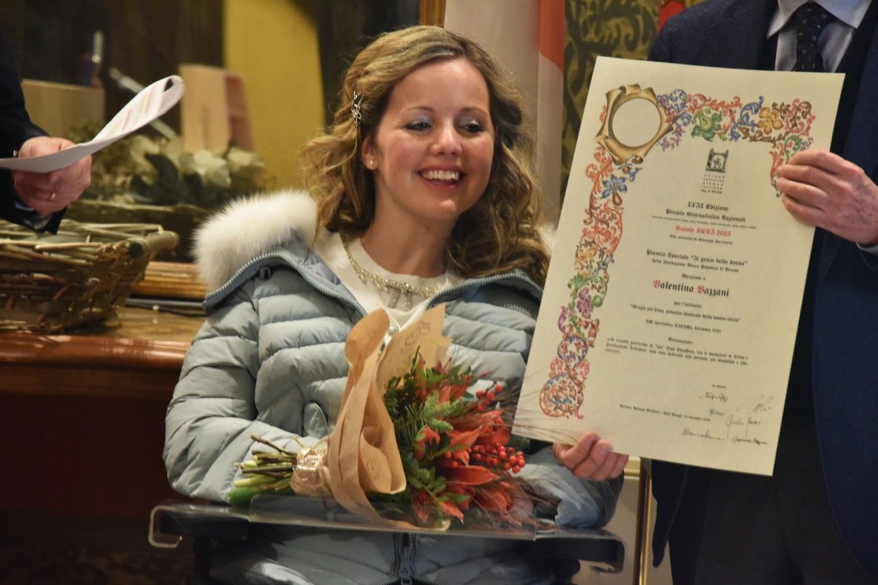 Una donna su sedia a rotelle tiene in mano un mazzo di fiori e un certificato.