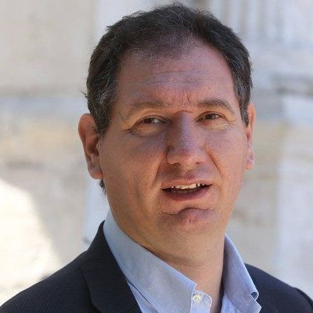 Un uomo in abito e camicia blu sorride alla telecamera.