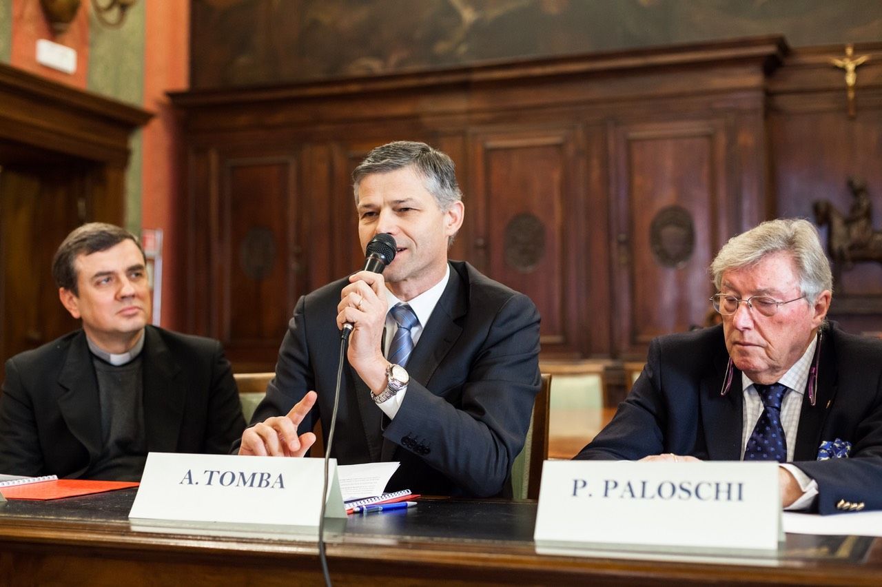 Un uomo parla al microfono mentre è seduto a un tavolo con altri due uomini.