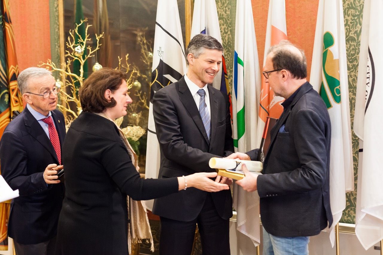 Un uomo in giacca e cravatta consegna un certificato a una donna davanti alle bandiere.