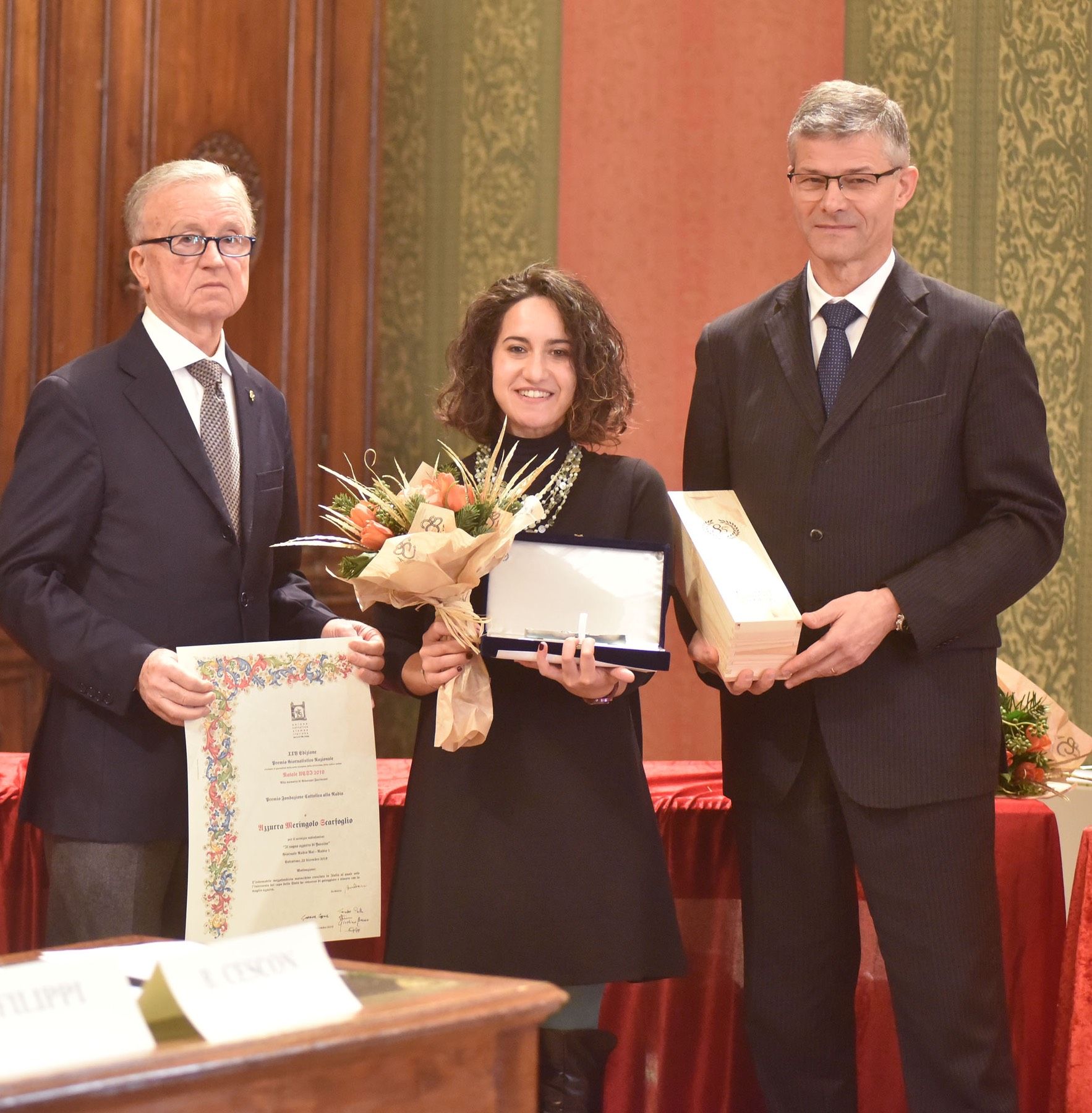 Una donna con in mano un mazzo di fiori si trova tra due uomini in giacca e cravatta
