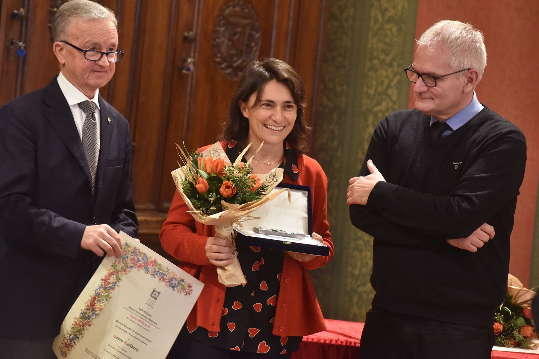 Una donna tiene in mano un mazzo di fiori e un certificato mentre due uomini guardano.