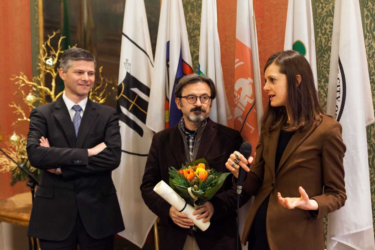 Un uomo con un cappello tiene in mano un mazzo di fiori mentre una donna parla al microfono.