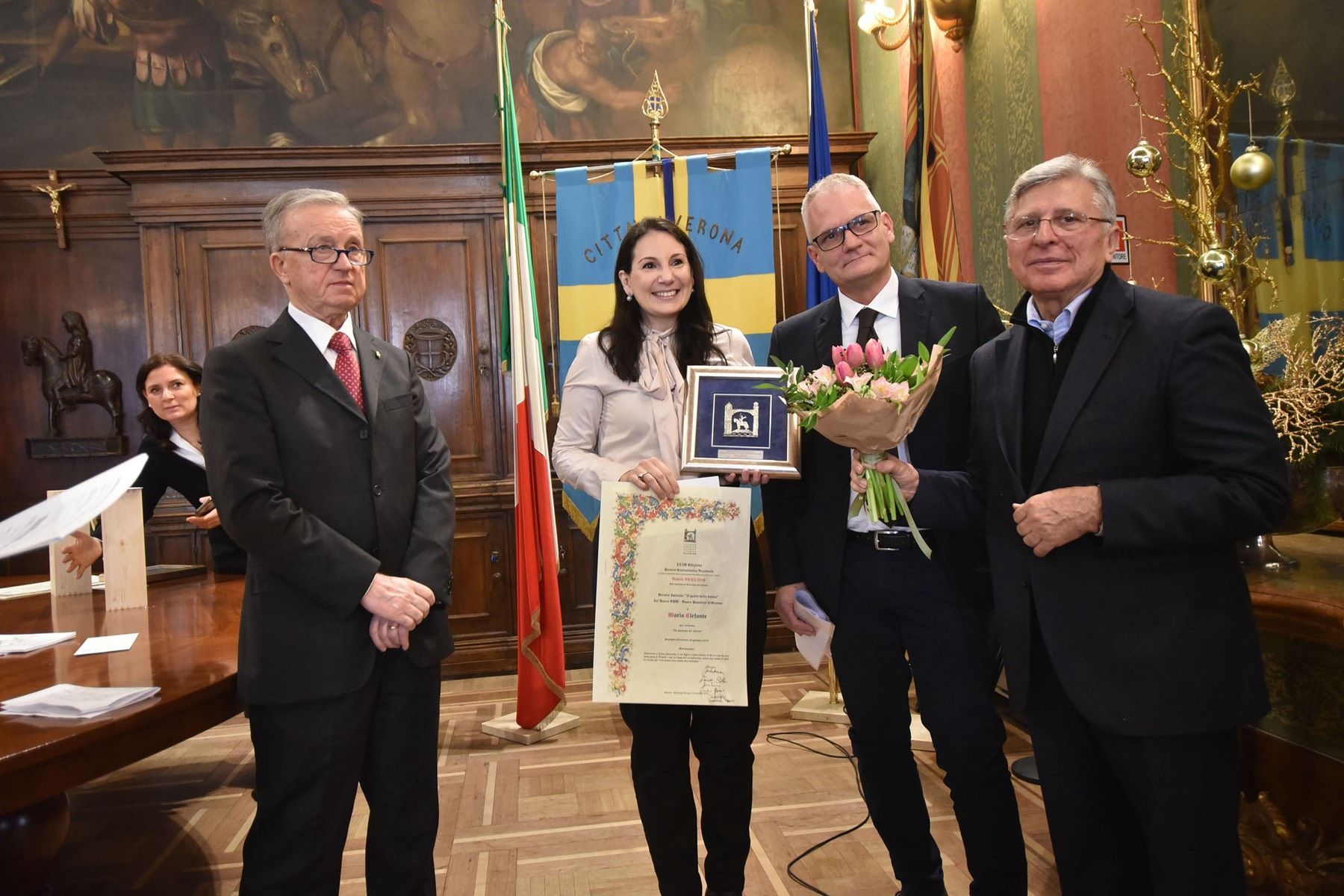 Una donna tiene in mano un certificato e un mazzo di fiori mentre sta in piedi accanto a tre uomini in giacca e cravatta.
