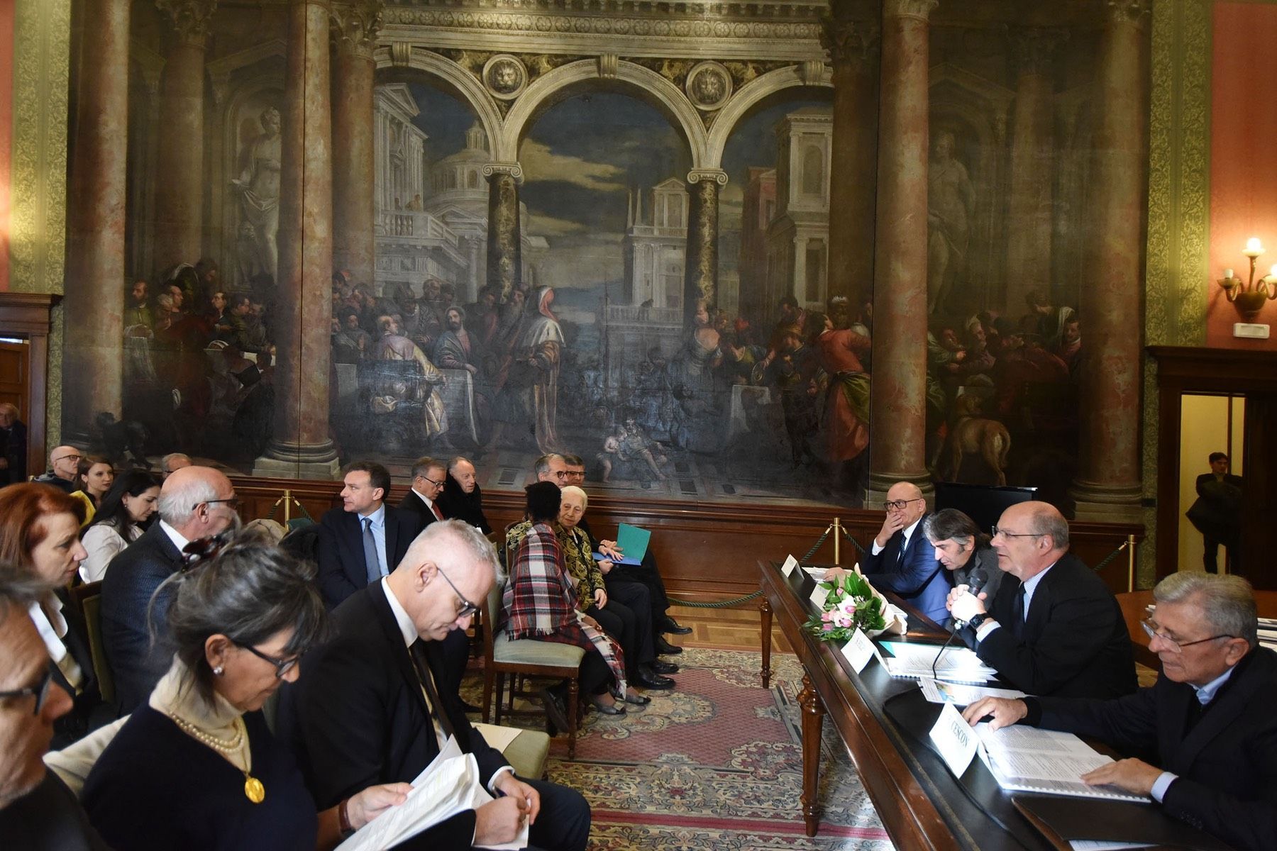 Un gruppo di persone è seduto ad un tavolo davanti ad un grande dipinto.