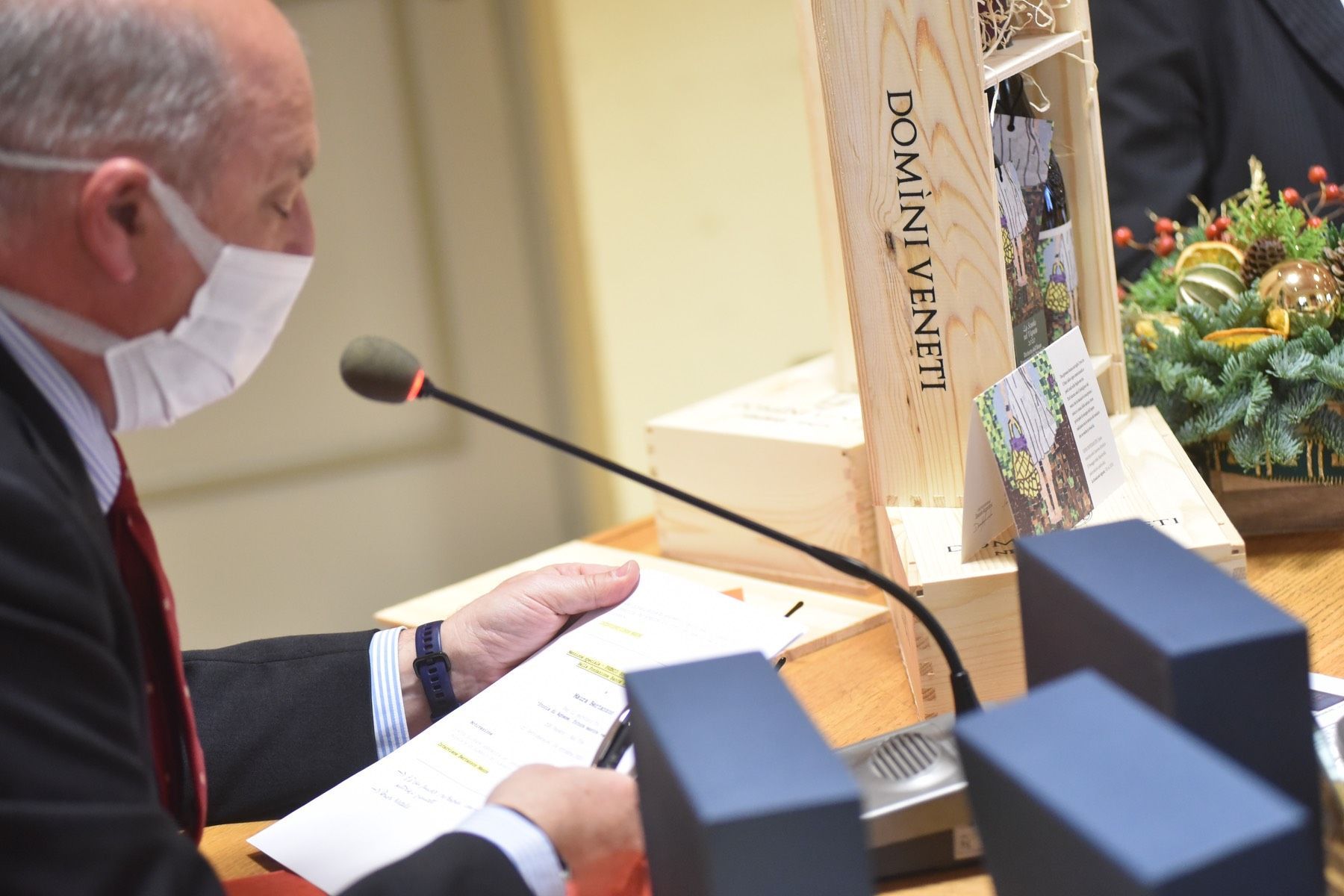 Un uomo che indossa una maschera è seduto a un tavolo e parla al microfono.