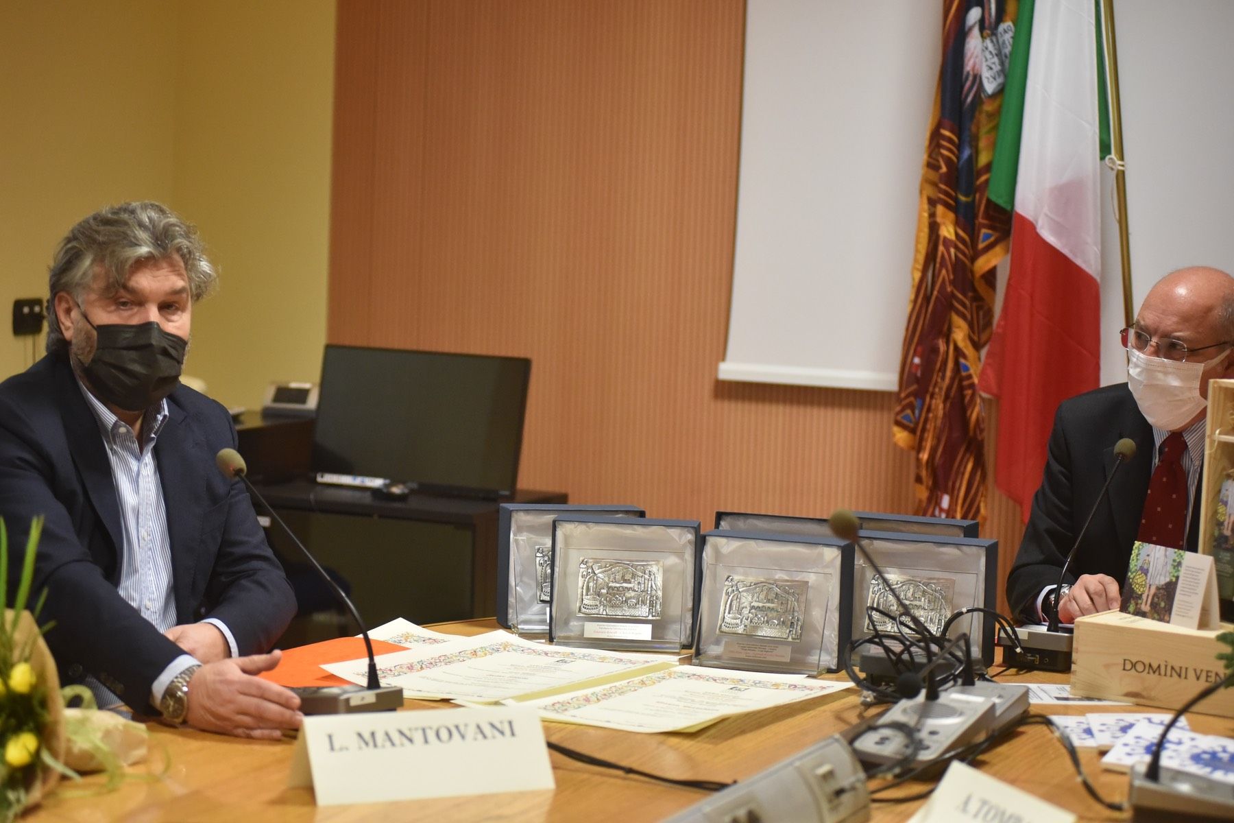 Due uomini che indossano maschere per il viso sono seduti a un tavolo con microfoni.