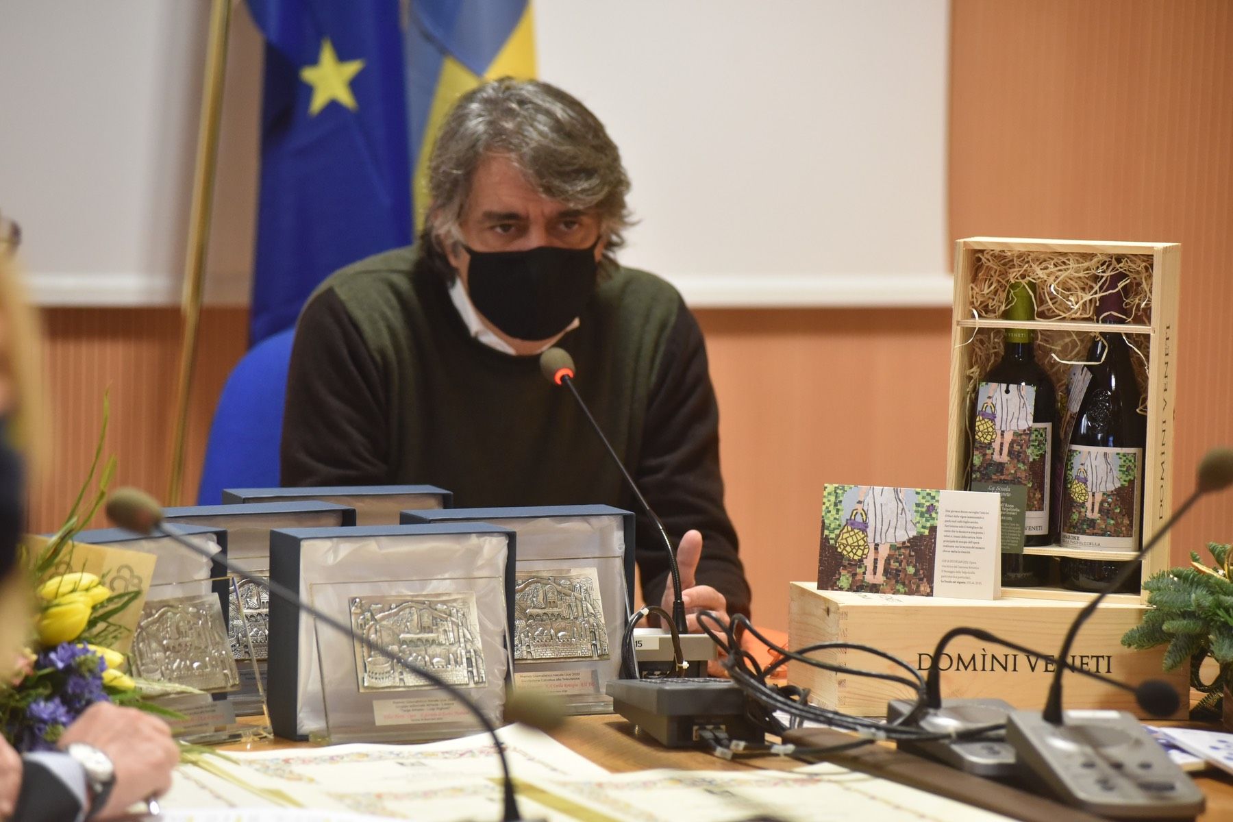 Un uomo che indossa una maschera è seduto a un tavolo con dei microfoni.