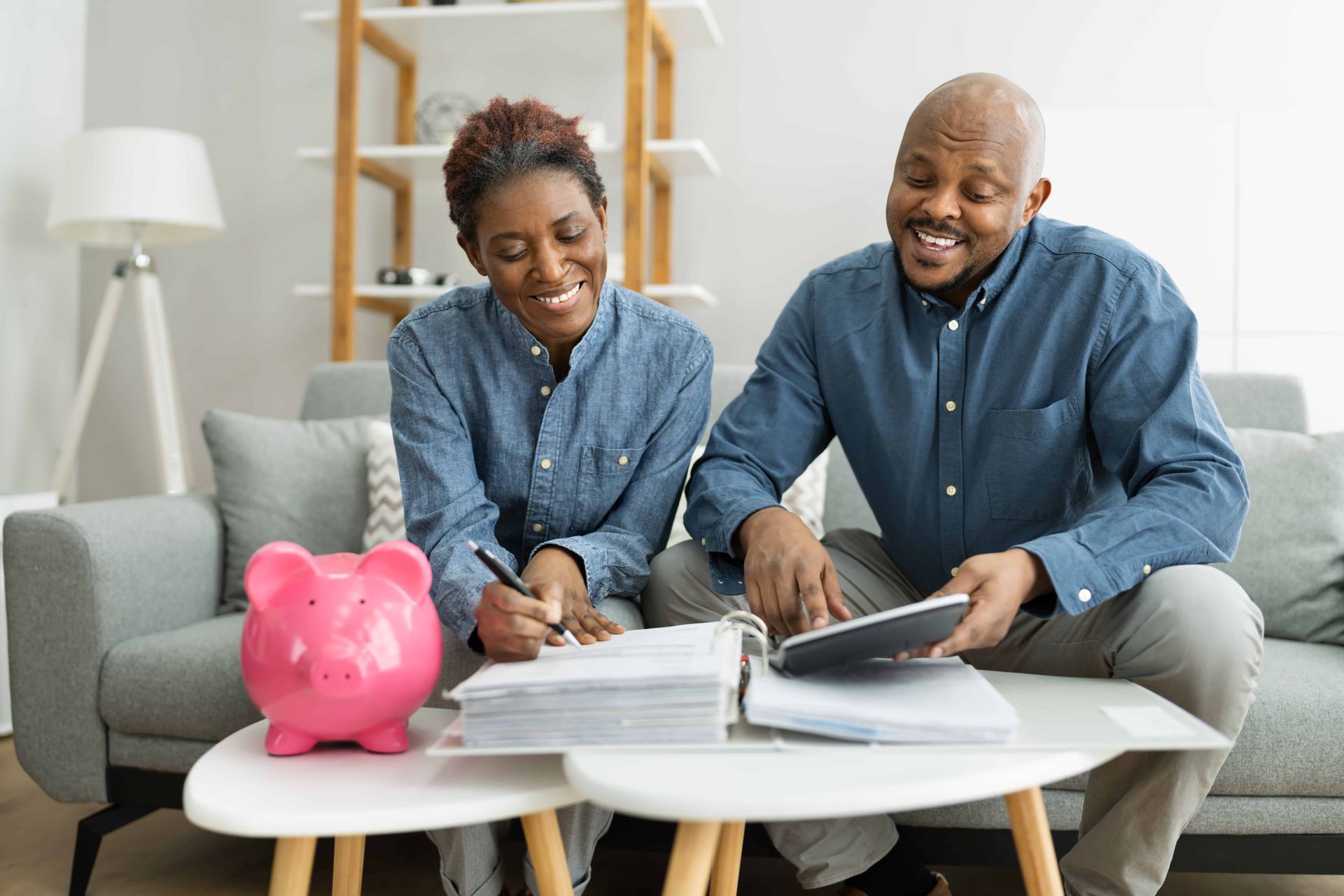 Couple reviews finances, smiling, with piggy bank, paperwork, and calculator on coffee table.